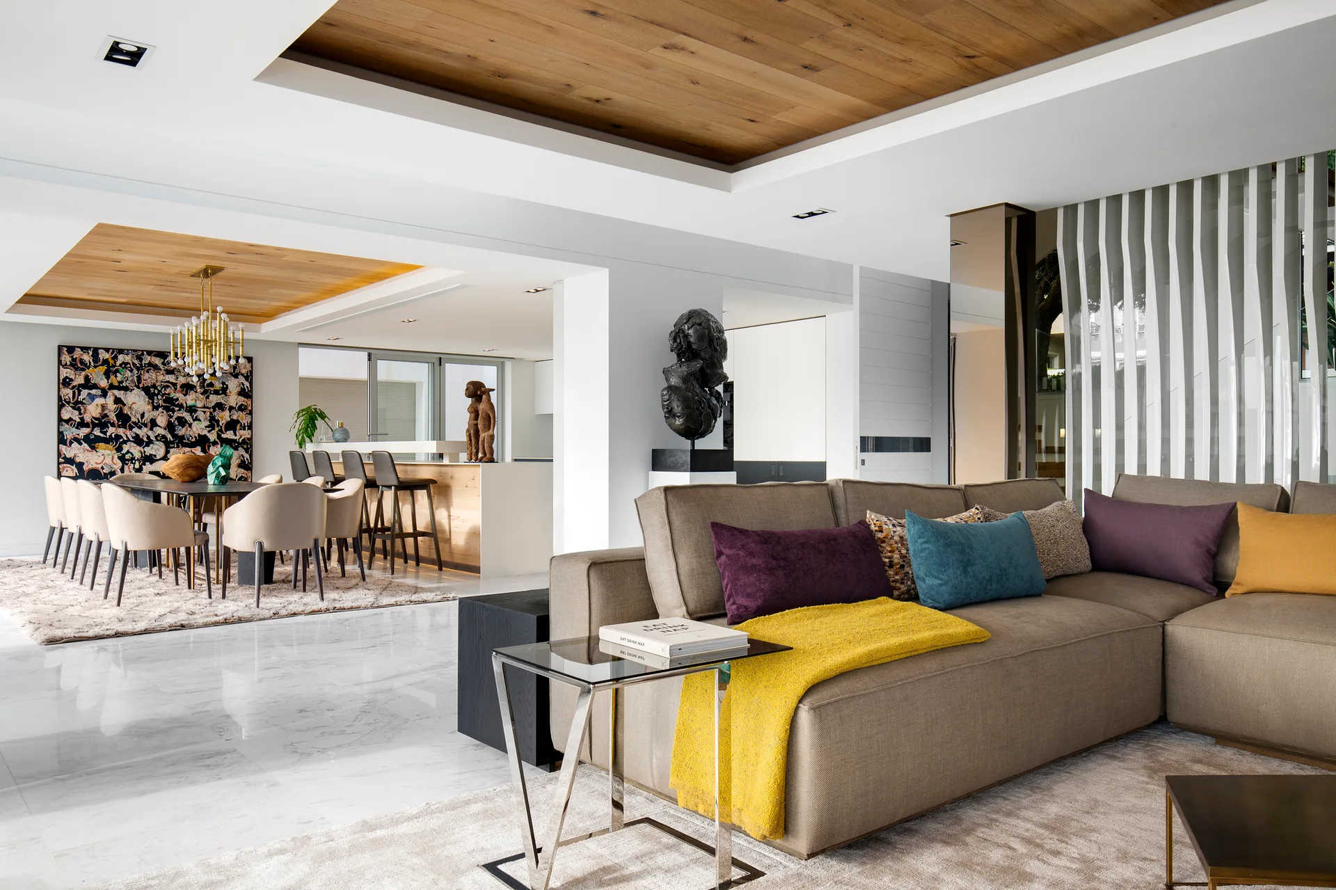 A modern open-plan living room features a tan sectional sofa with colorful pillows, polished white marble flooring, and recessed wood ceiling panels.