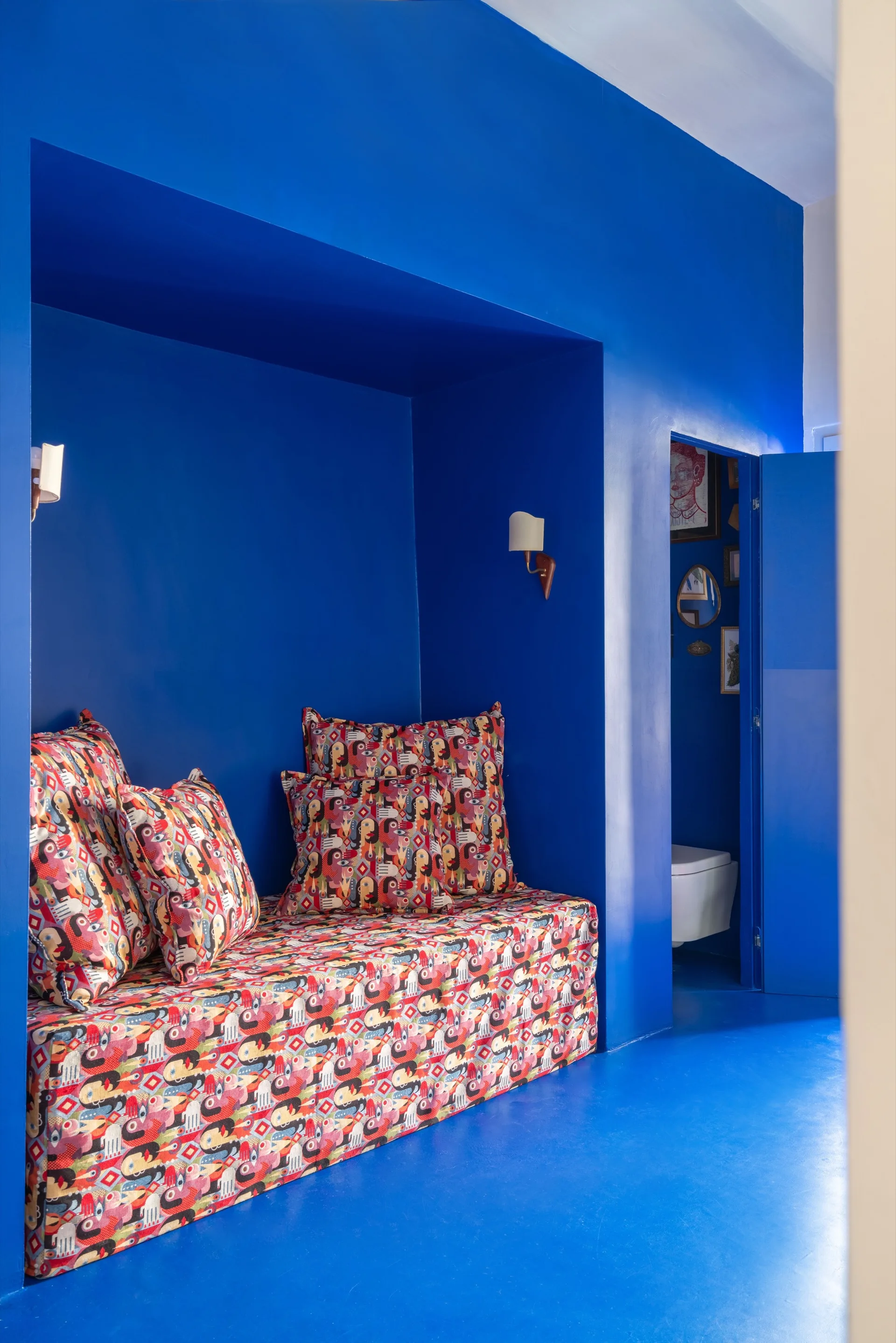 A built-in daybed with colorful geometric pattern upholstery sits inside a recessed niche with solid blue painted walls and floors.