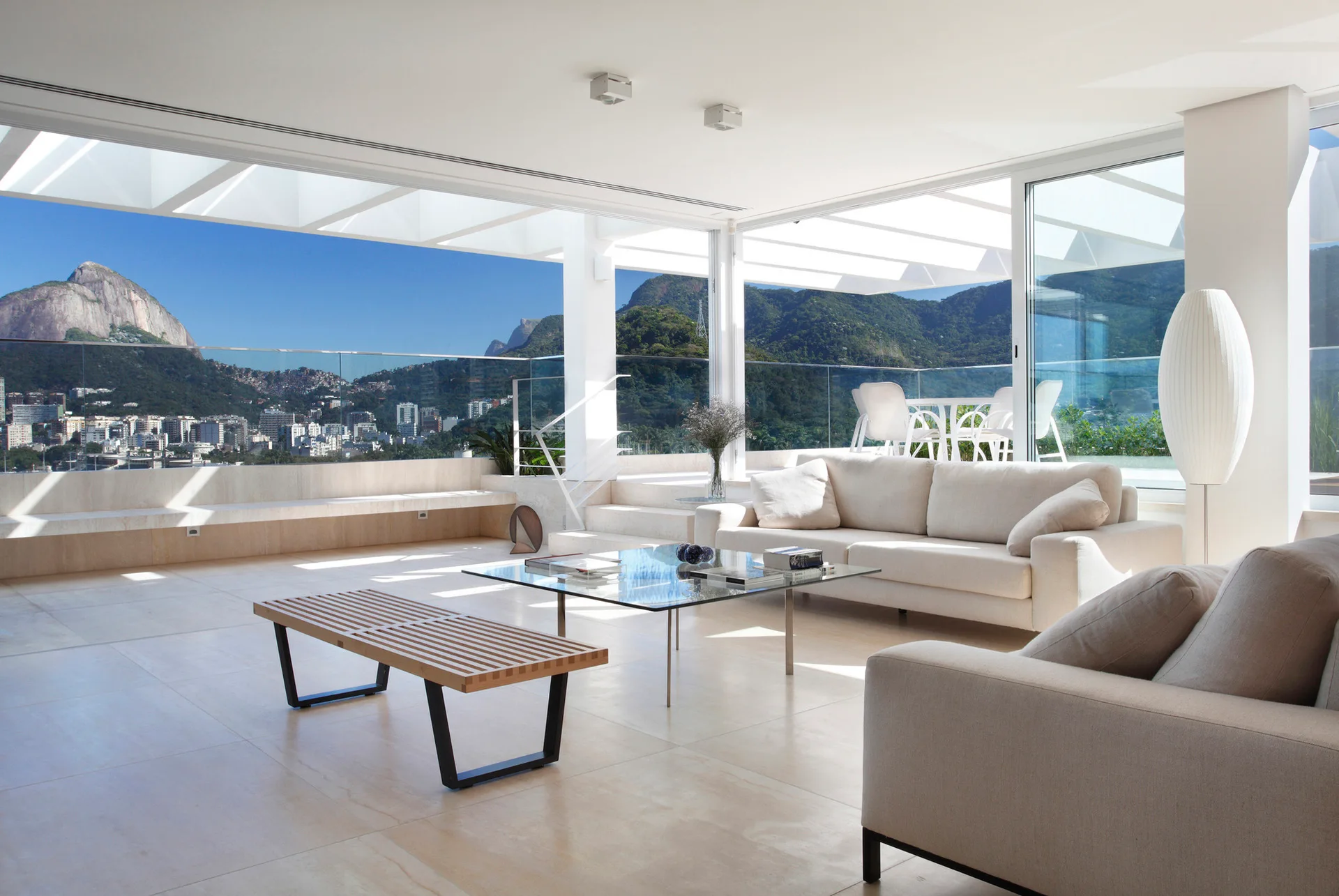 A bright, minimalist living room with cream sofas, a glass coffee table, and large sliding glass doors overlooking mountains.