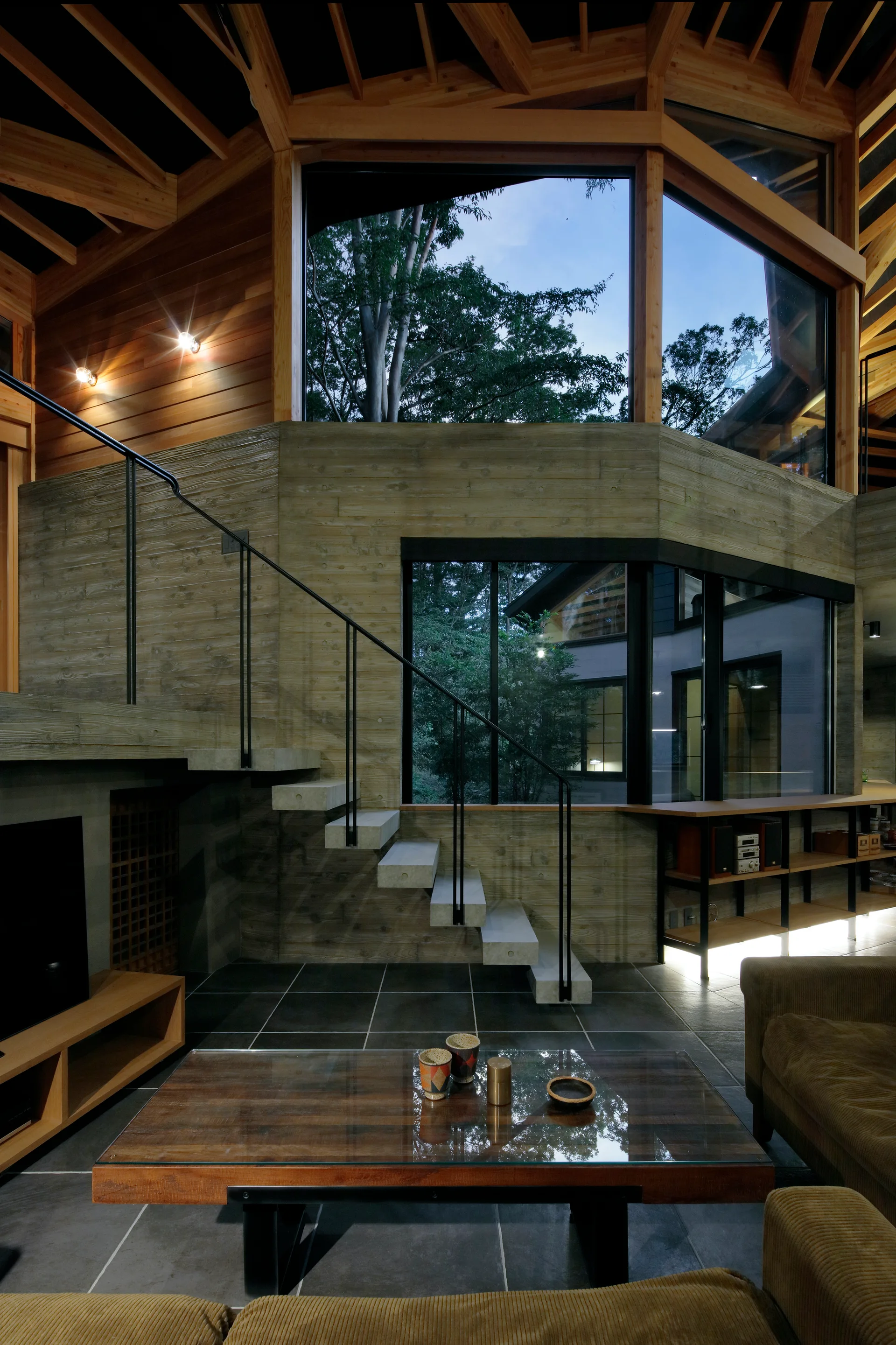 A modernist living room features a floating concrete staircase, board-formed concrete walls, large windows, and a geometric timber ceiling.