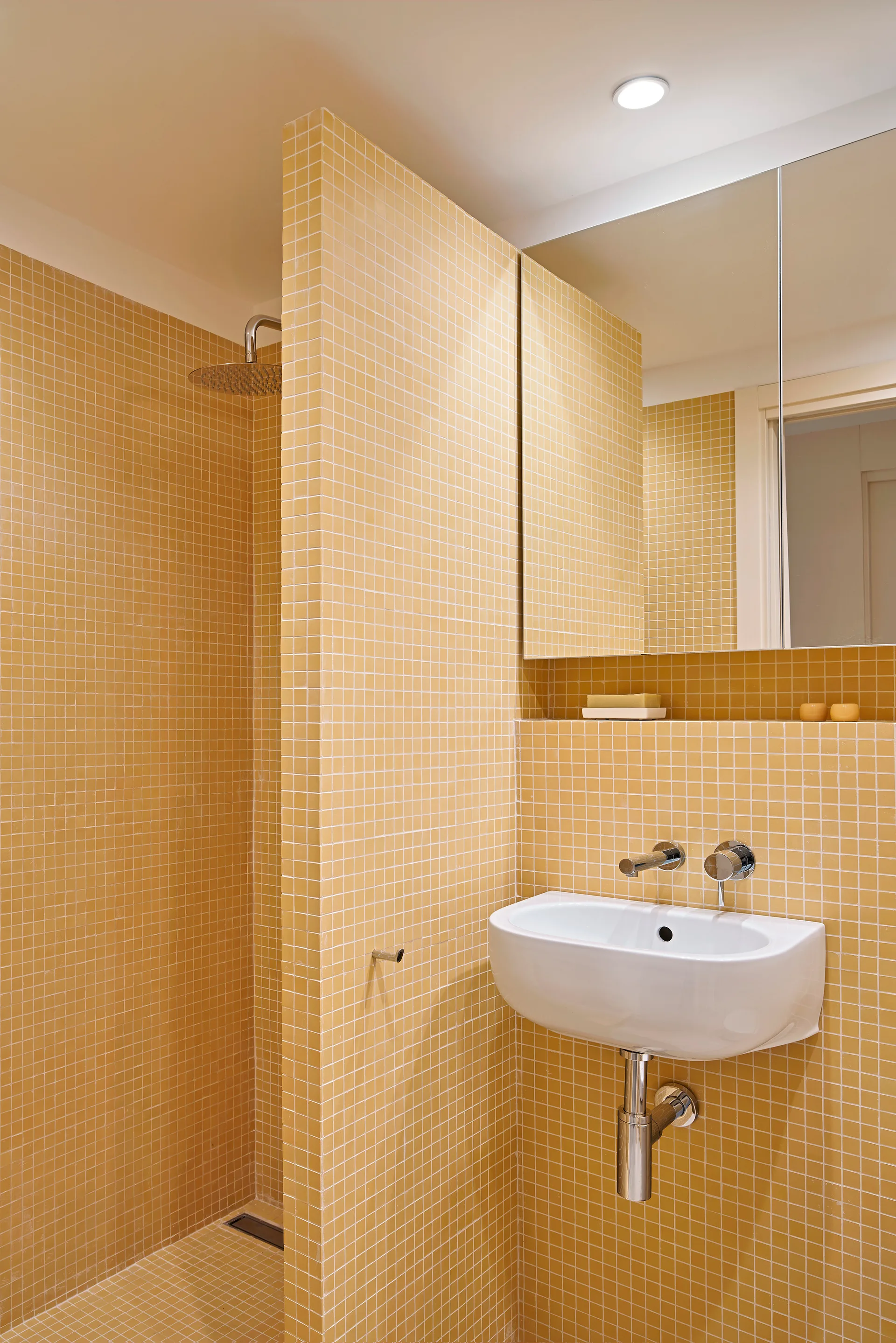 A minimalist bathroom features yellow grid mosaic tiles on the walls and floor, a white wall-mounted washbasin, and a recessed storage shelf.