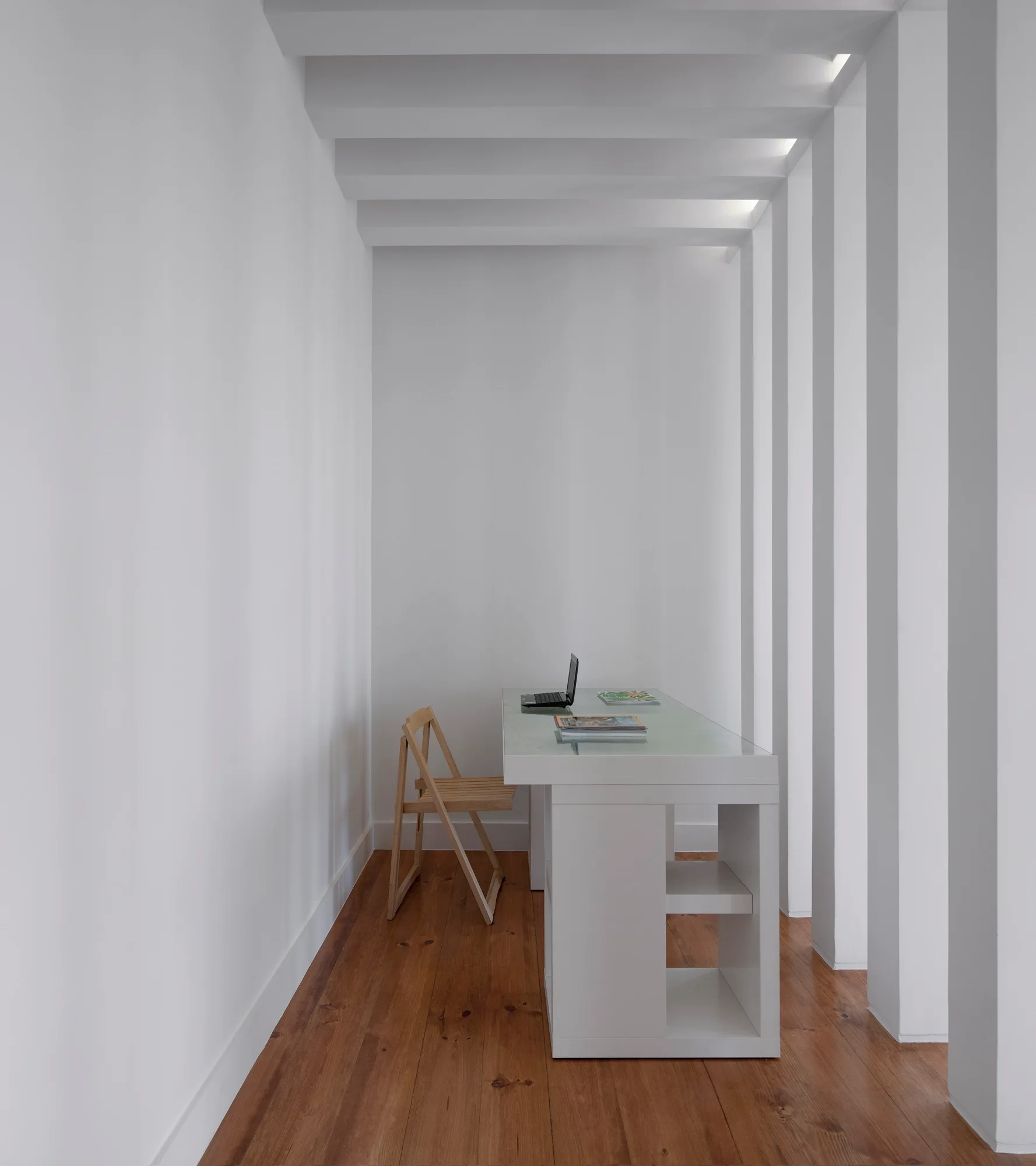A minimalist home office features a white desk, a wooden folding chair, medium-toned wood floors, and a wall of vertical architectural fins.