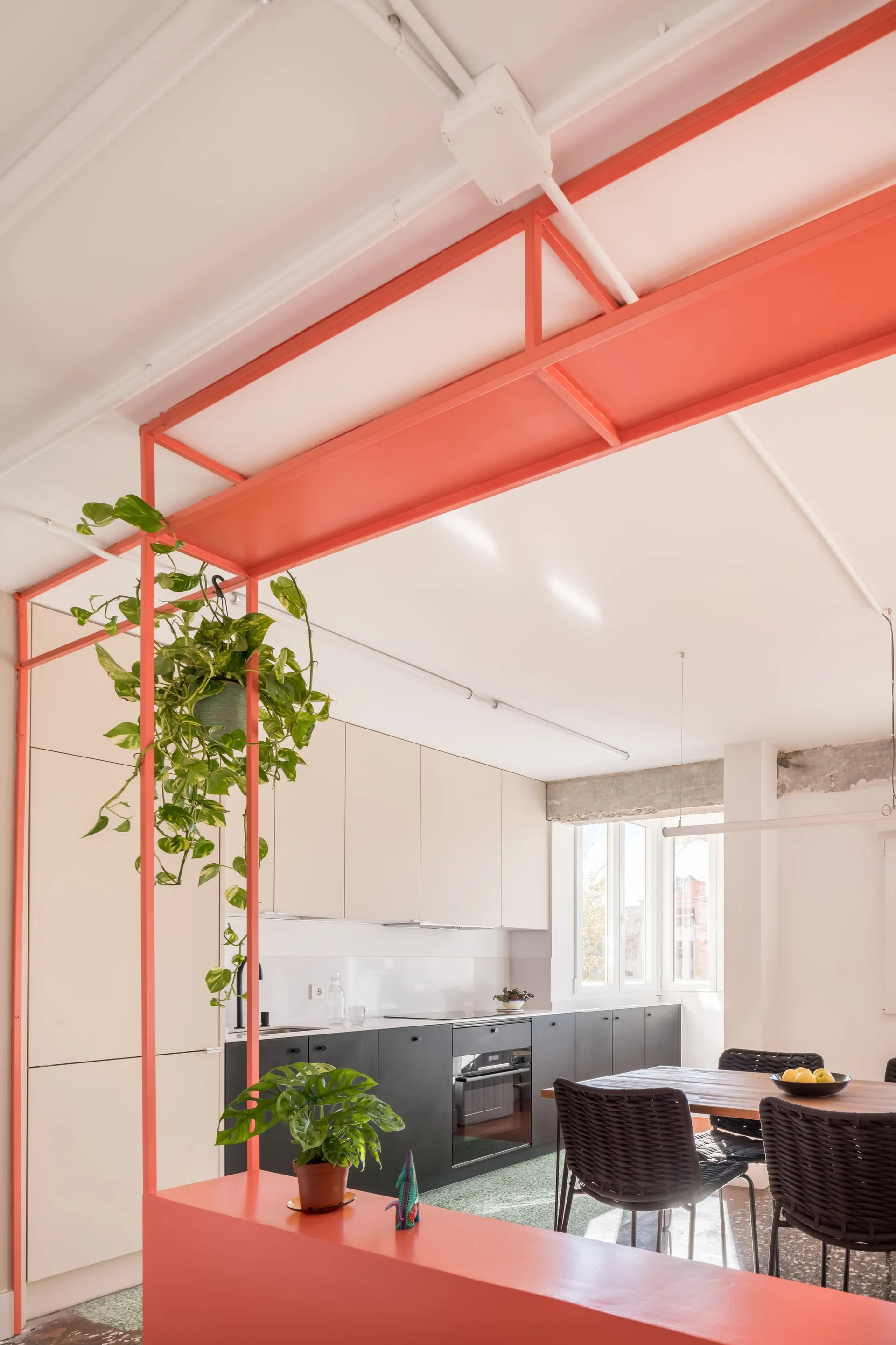 An open kitchen and dining area features an orange-red metal structural frame holding a hanging plant, two-tone cabinets, and a wooden dining table.