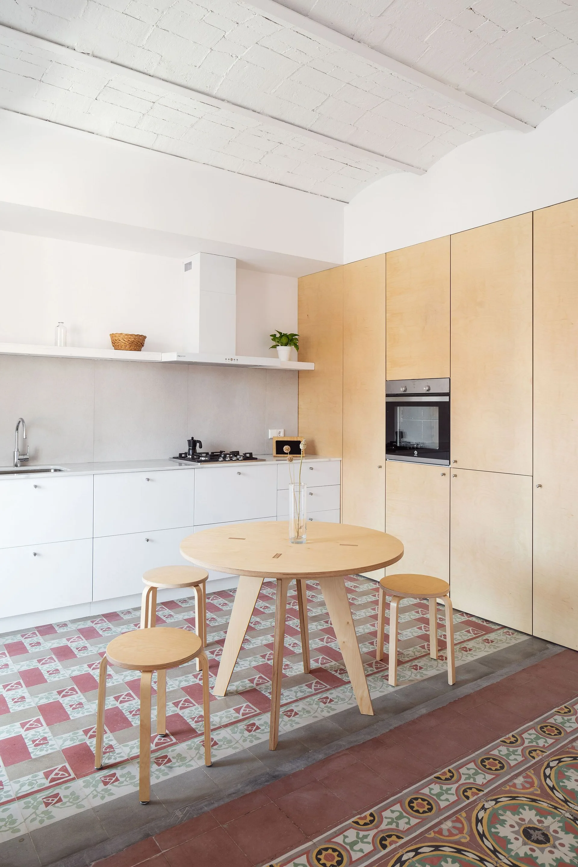 A kitchen featuring a round wood dining table with stools under a white vaulted brick ceiling, with white and light wood cabinets and patterned tile floors.