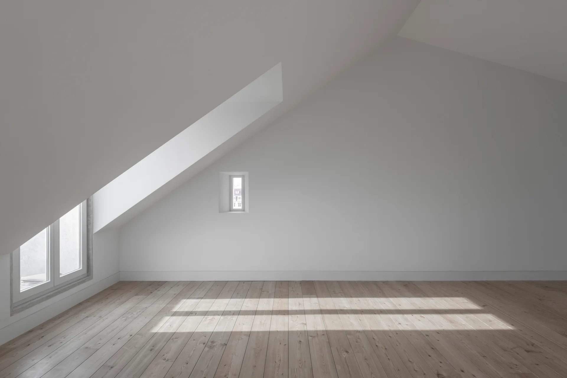 An empty attic room featuring white sloped walls, a light wood plank floor, and natural sunlight streaming from two windows.