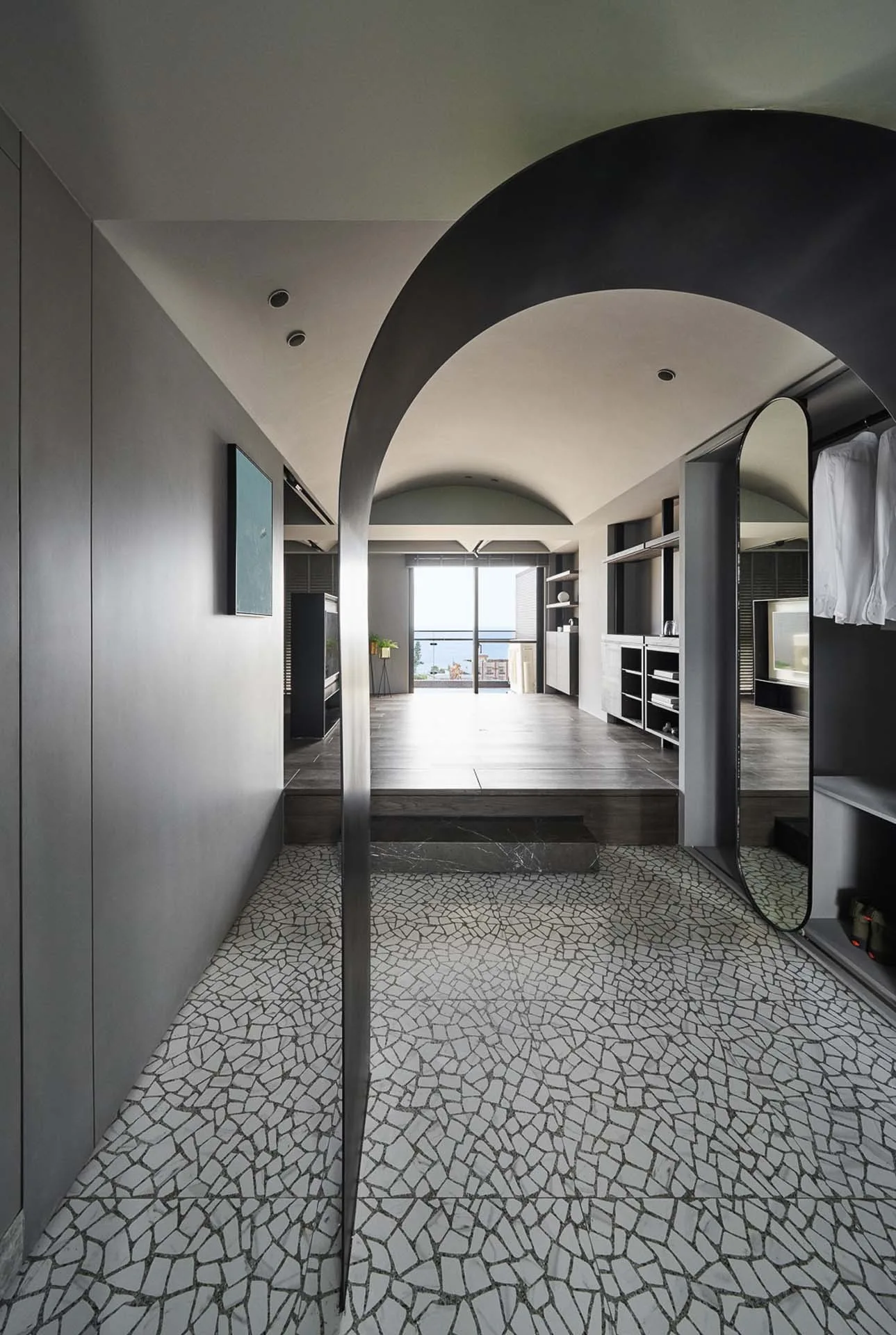 A modern entryway with a grey stone mosaic tile floor and a dark arched entryway leading to a raised wood-floor bedroom.
