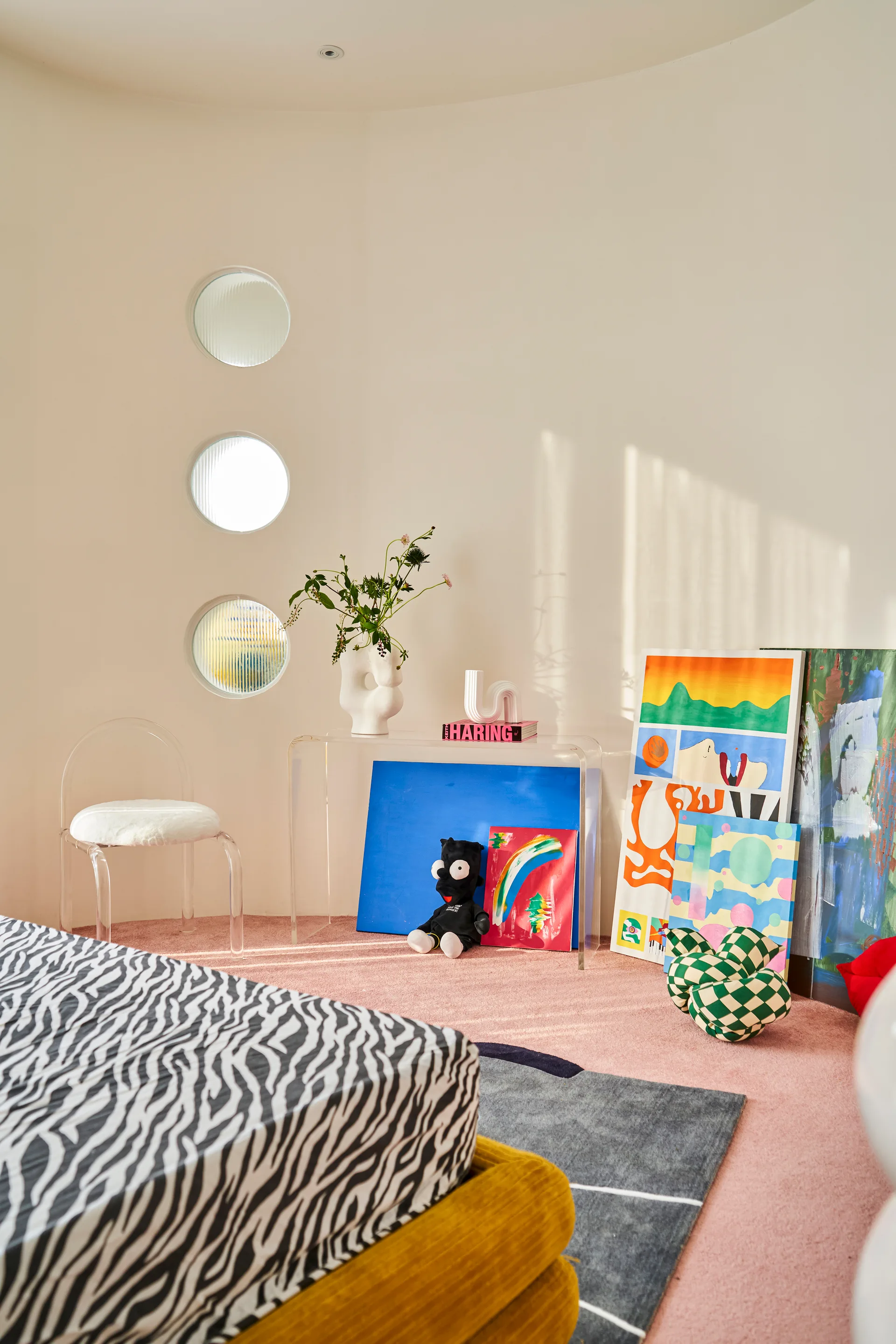 A bedroom features a curved cream wall with three stacked circular windows, a clear acrylic console table, and a bed with zebra print bedding.