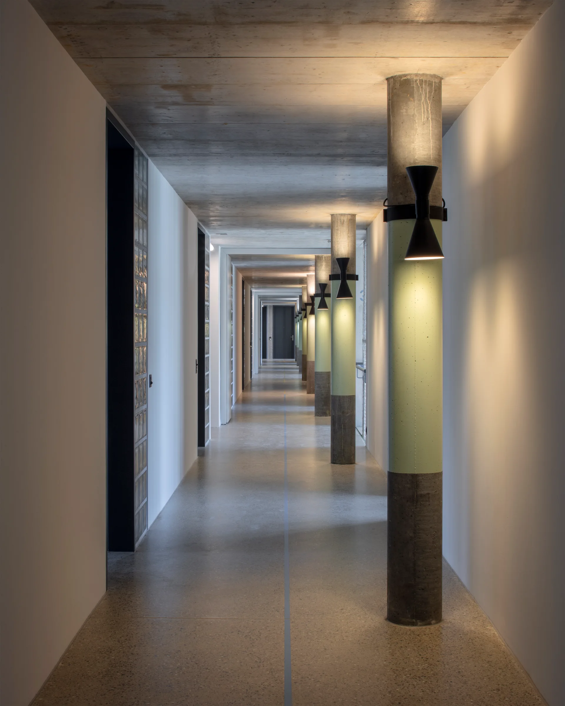 A long minimalist corridor features concrete columns painted sage green and black wall sconces beneath a raw concrete ceiling.