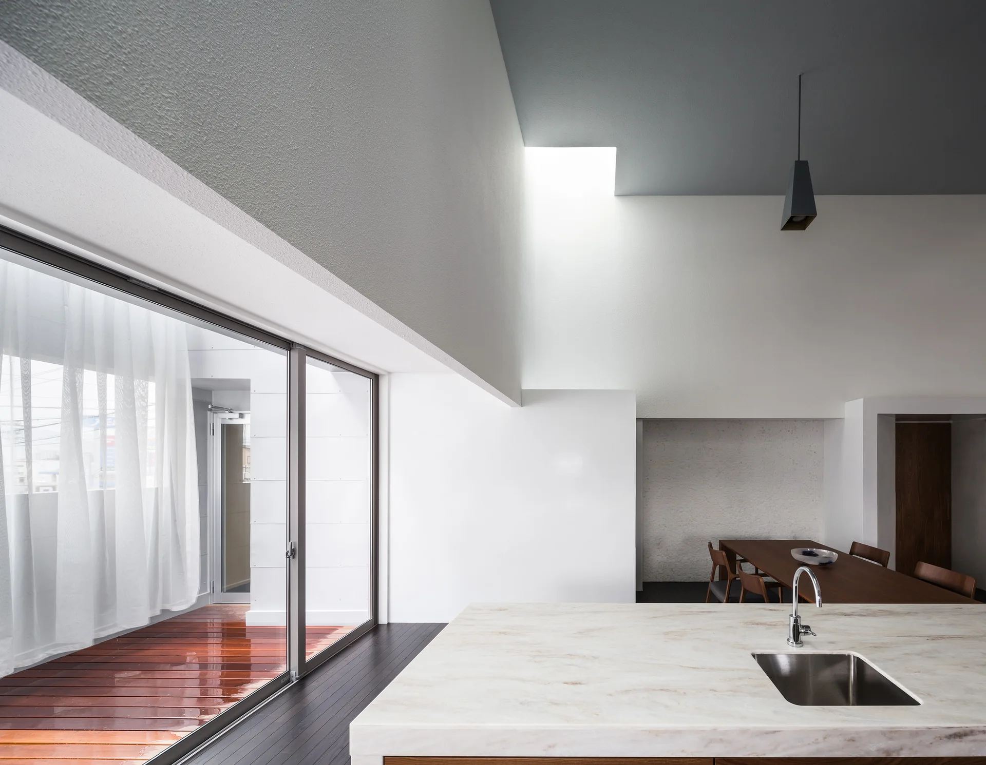 A minimalist kitchen features a marble-topped island with a sink, a dining table in the background, and sliding doors opening to a wood deck.
