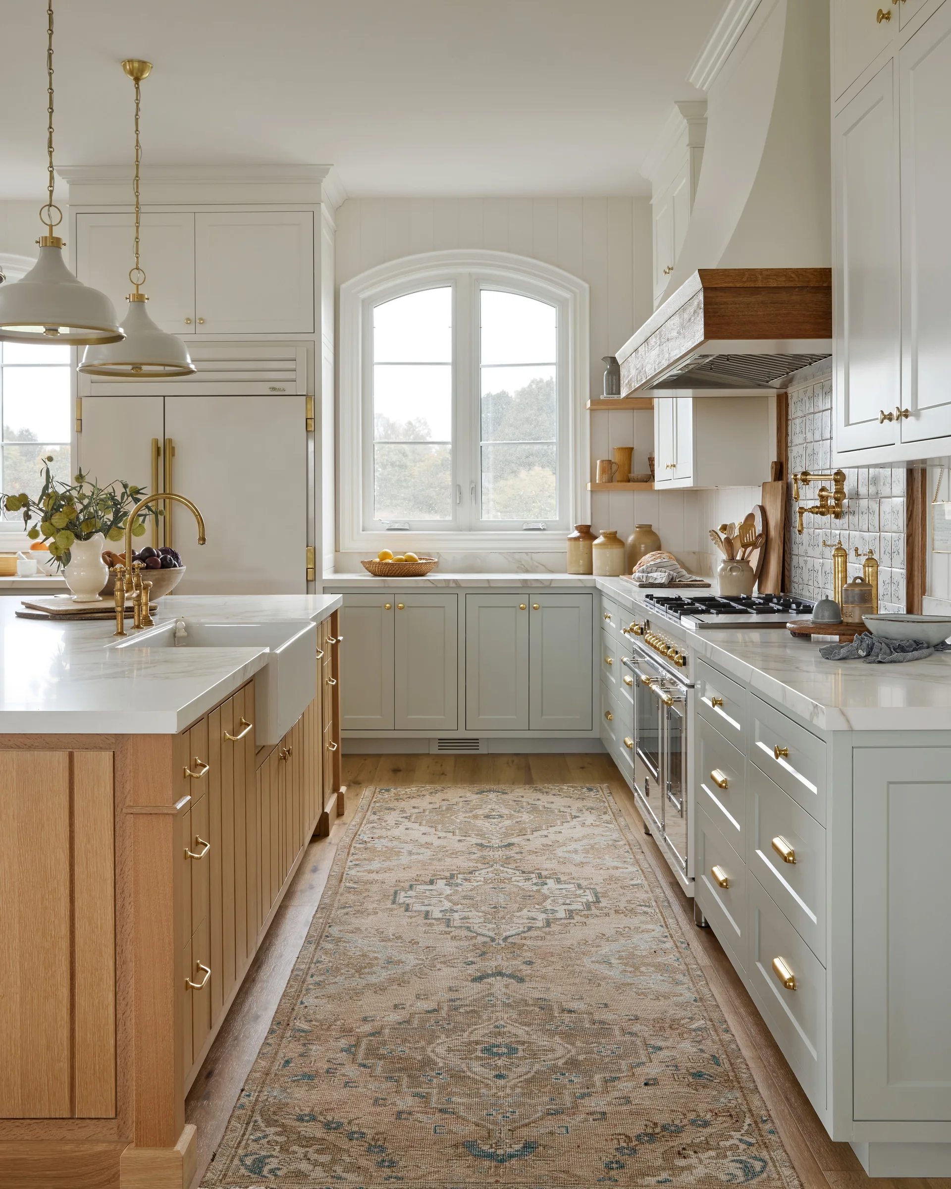 A kitchen features a light oak island with a farmhouse sink, light gray cabinets, brass hardware, and a patterned runner rug.