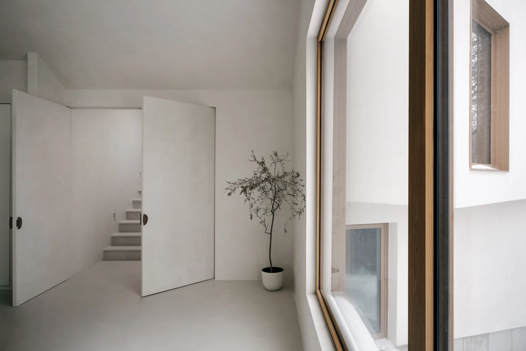 A minimalist white hallway features two large double doors open to a stone staircase next to a large window with thick wood trim.