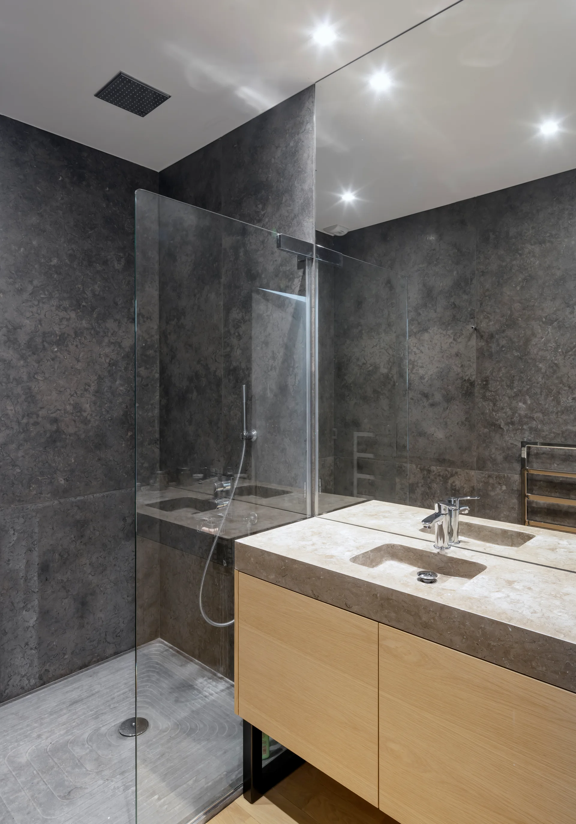 A modern bathroom showcases a light wood vanity with an integrated stone sink set against dark grey tiled walls and a glass shower panel.