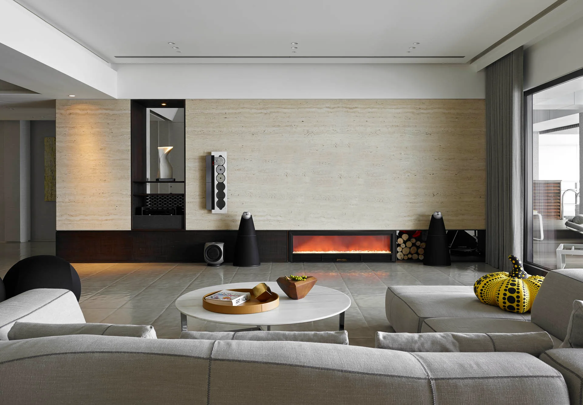 A modern living room features a large beige stone accent wall with an integrated linear fireplace, a grey sectional sofa, and a marble coffee table.