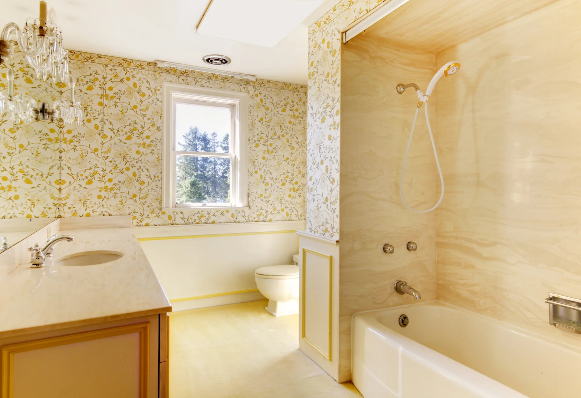A traditional bathroom features yellow floral wallpaper, a white bathtub with a beige stone shower surround, and a wooden vanity.
