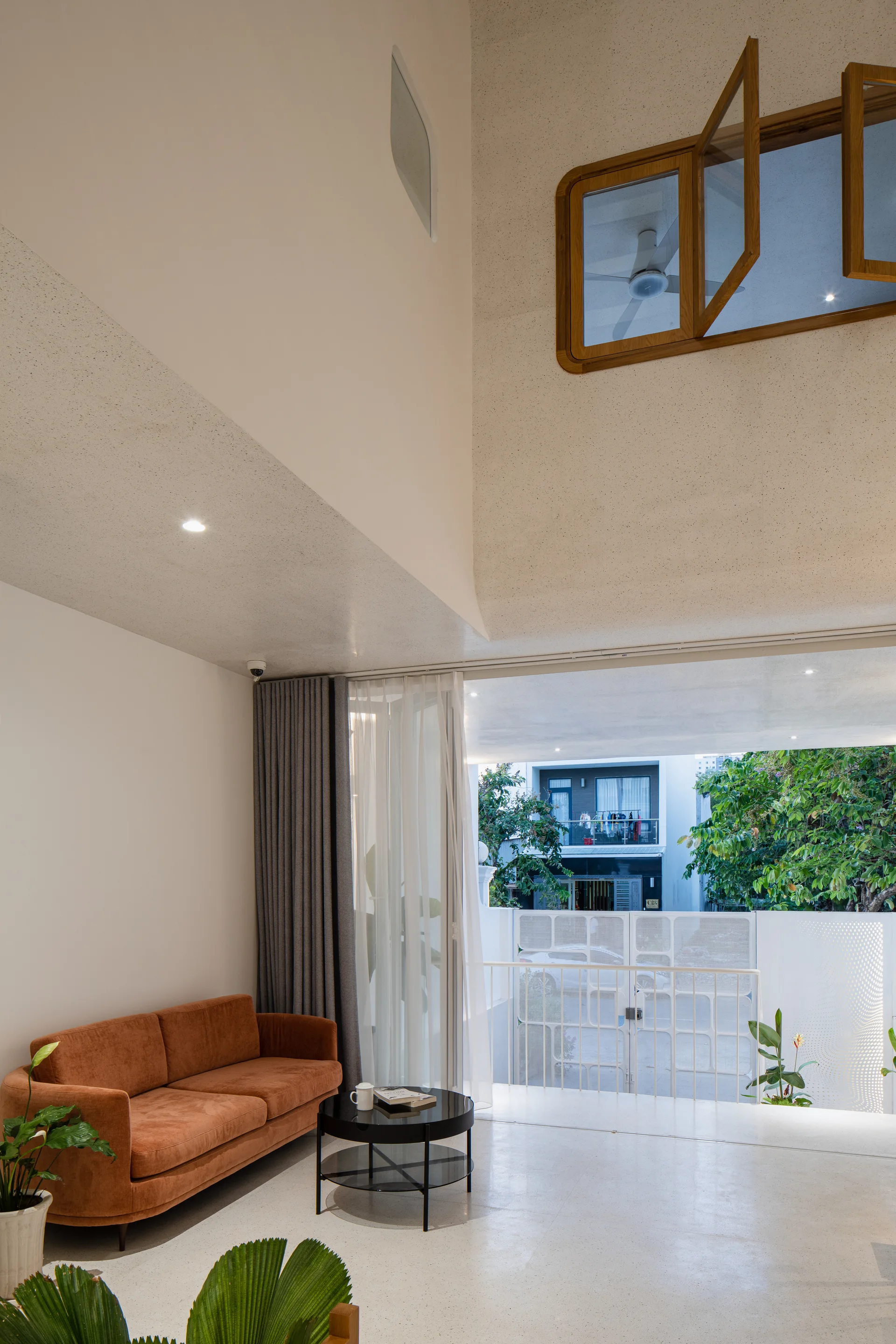A modern double-height living room features a burnt orange sofa, a black coffee table, speckled terrazzo flooring, and a balcony railing.