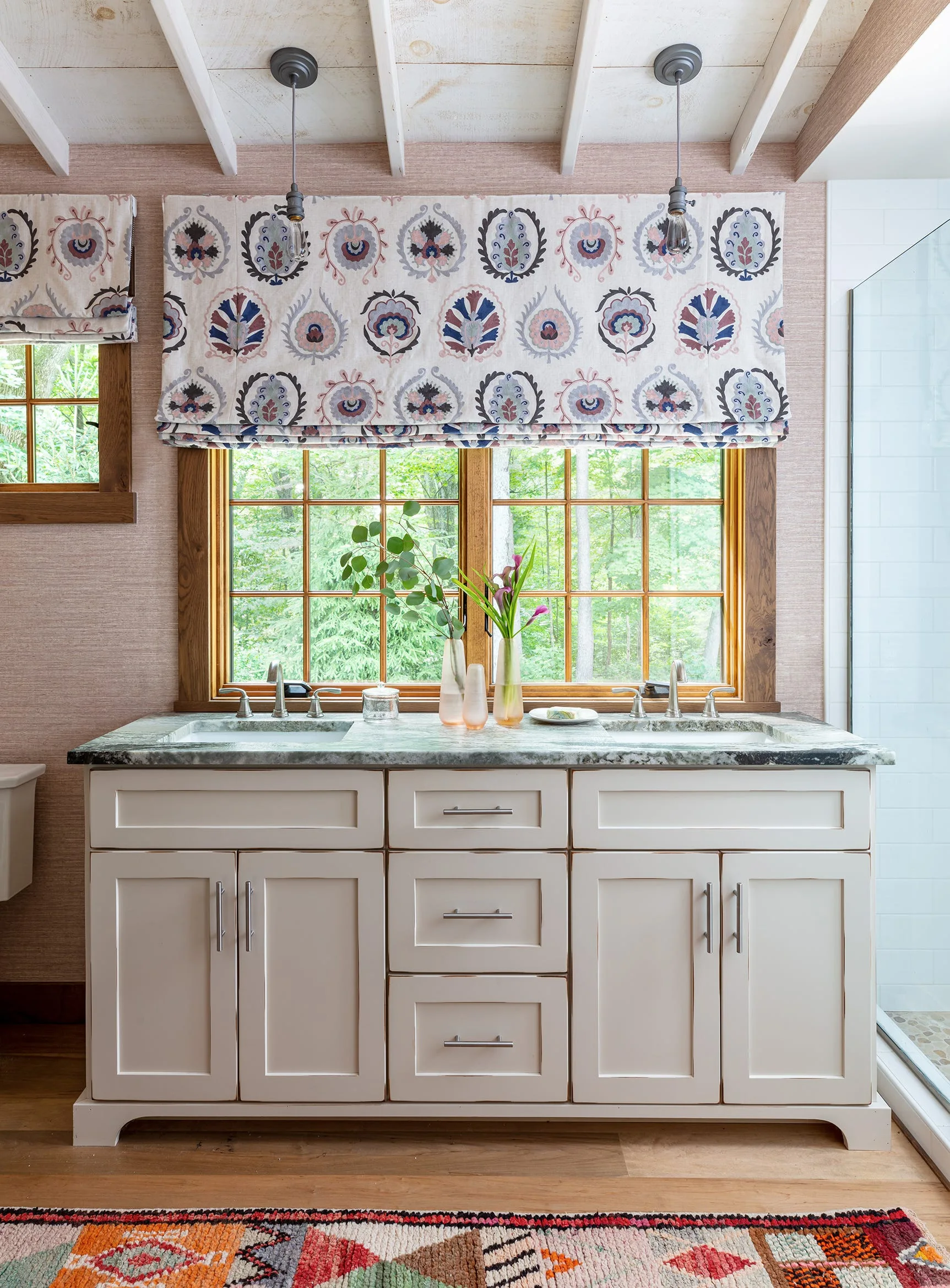 A cream double-sink vanity with a green stone countertop is positioned in front of a wood-framed window under a patterned Ikat Roman shade.