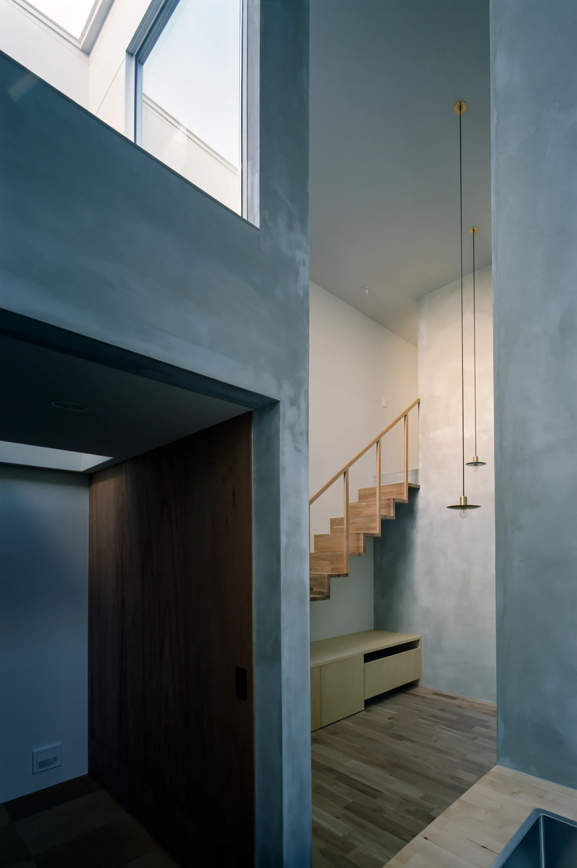 A minimalist wooden staircase stands against a tall grey concrete wall with two slender brass pendant lights hanging from the high ceiling.