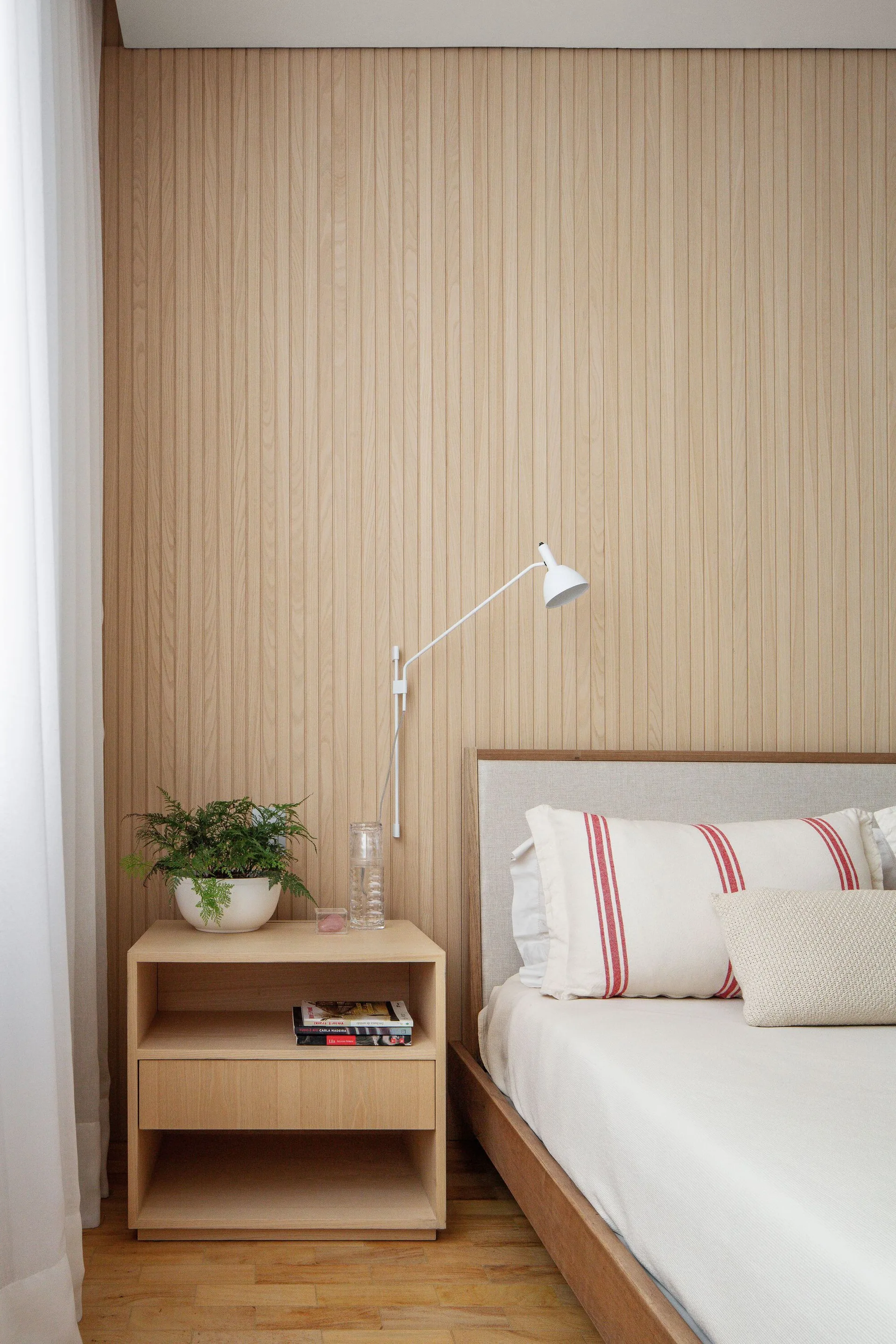 A minimalist bedroom corner features vertical slatted wood wall paneling, a wooden bed with striped pillows, and an open-shelf nightstand.