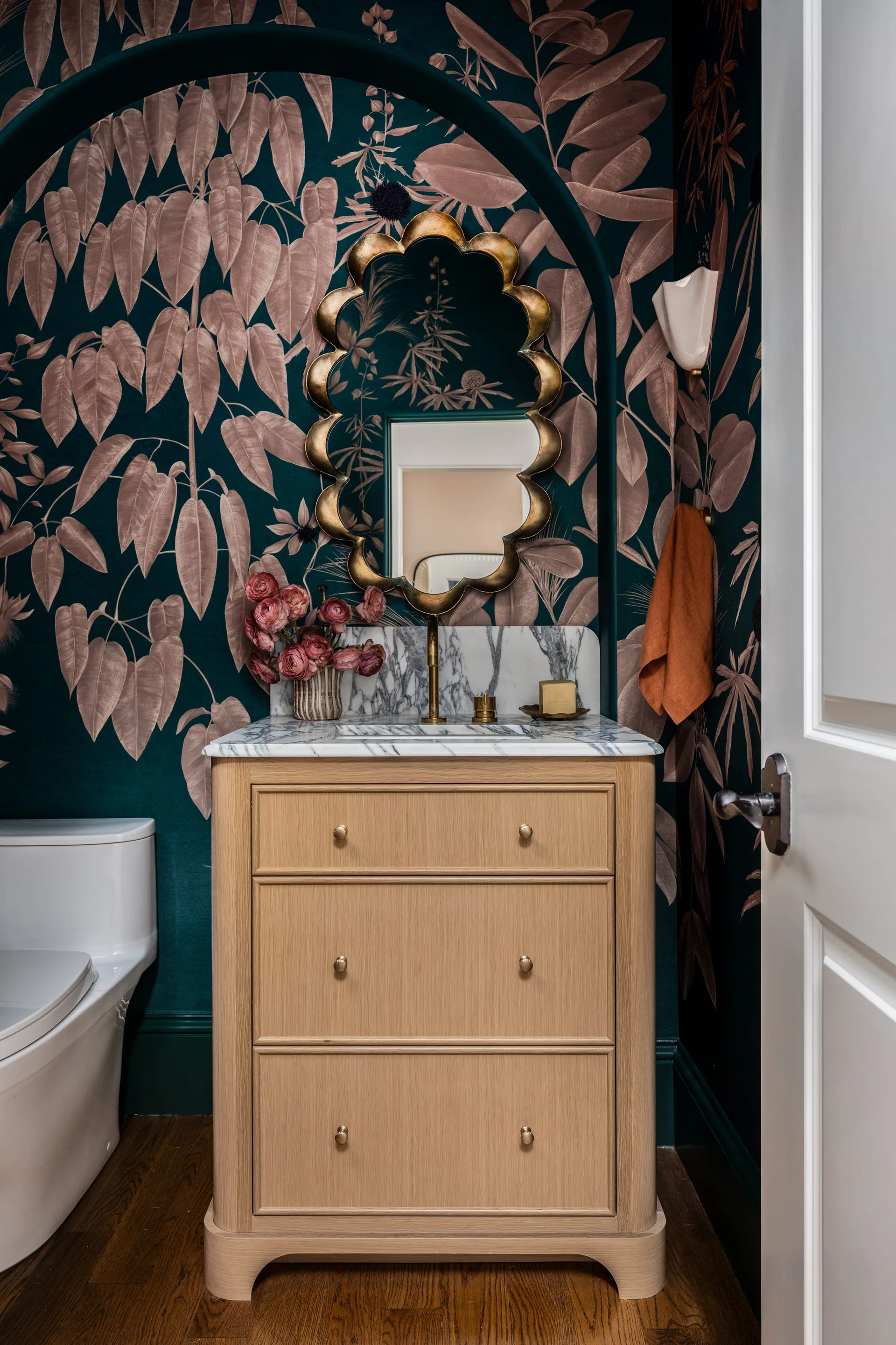 A light oak vanity with a marble countertop stands against dark green botanical wallpaper under a scalloped gold mirror in a powder room.