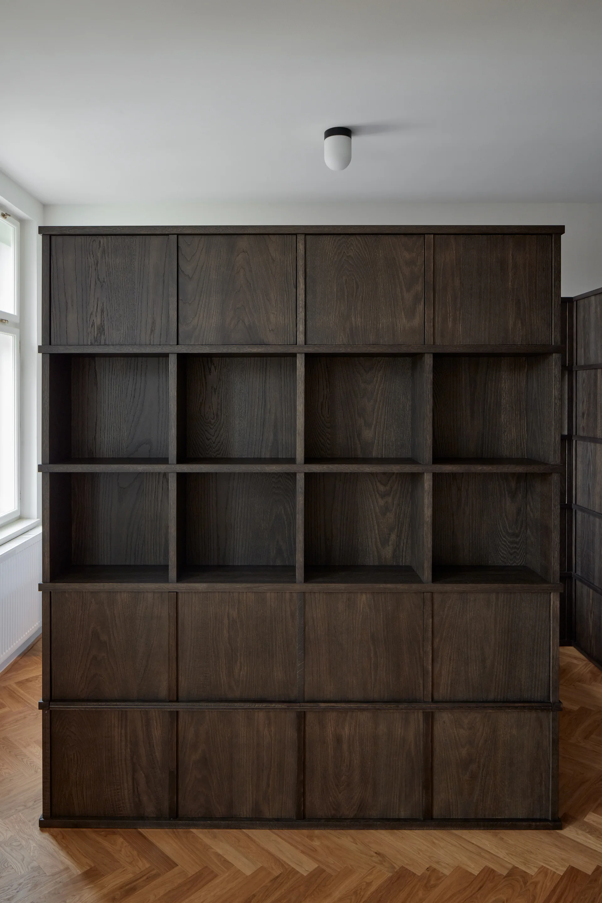 A large dark-stained wood grid shelving unit stands on a light oak herringbone parquet floor against a plain white wall.