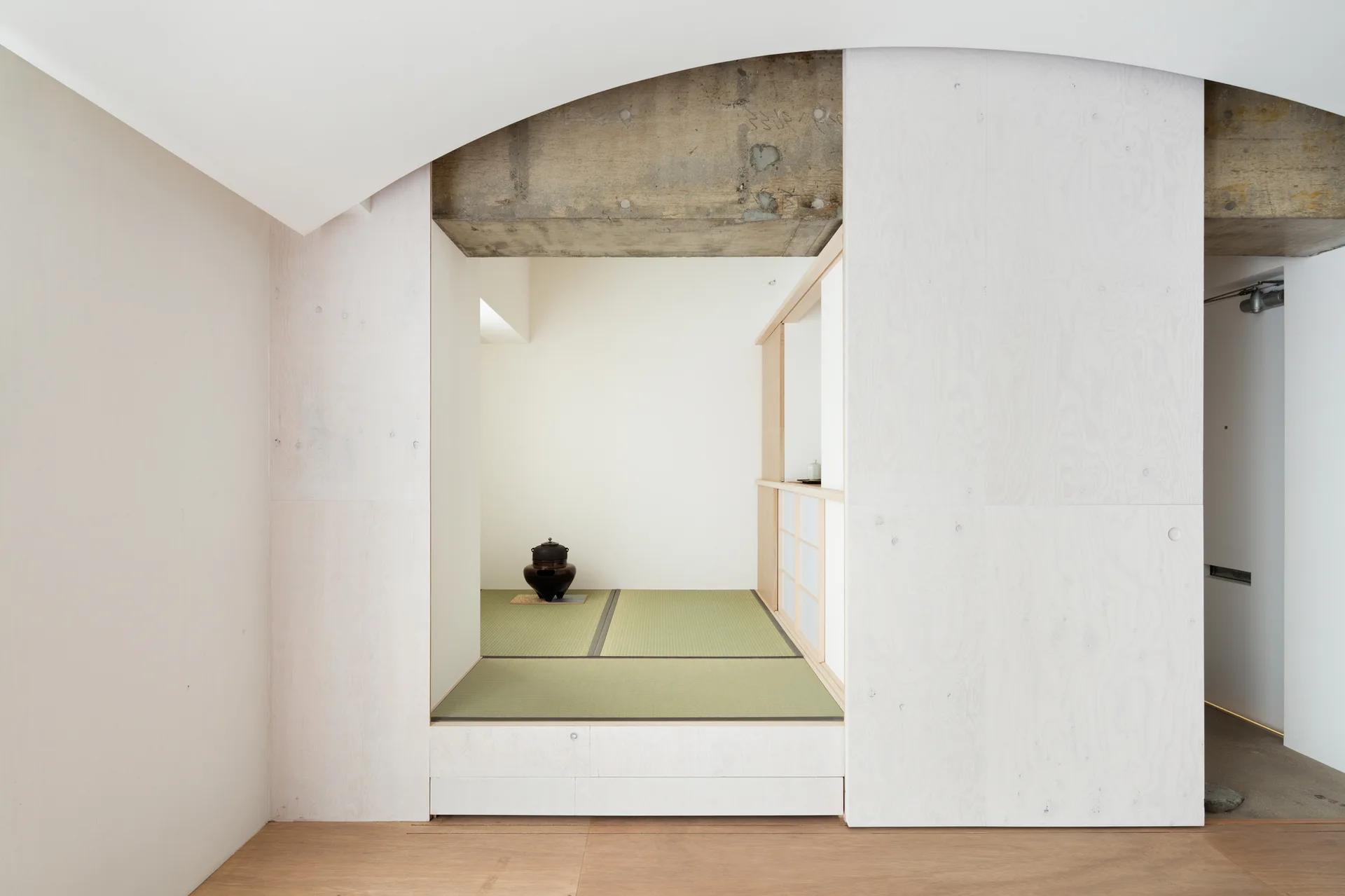 A minimalist interior space with a raised tatami floor area, light wood paneled walls, and a raw exposed concrete beam overhead.