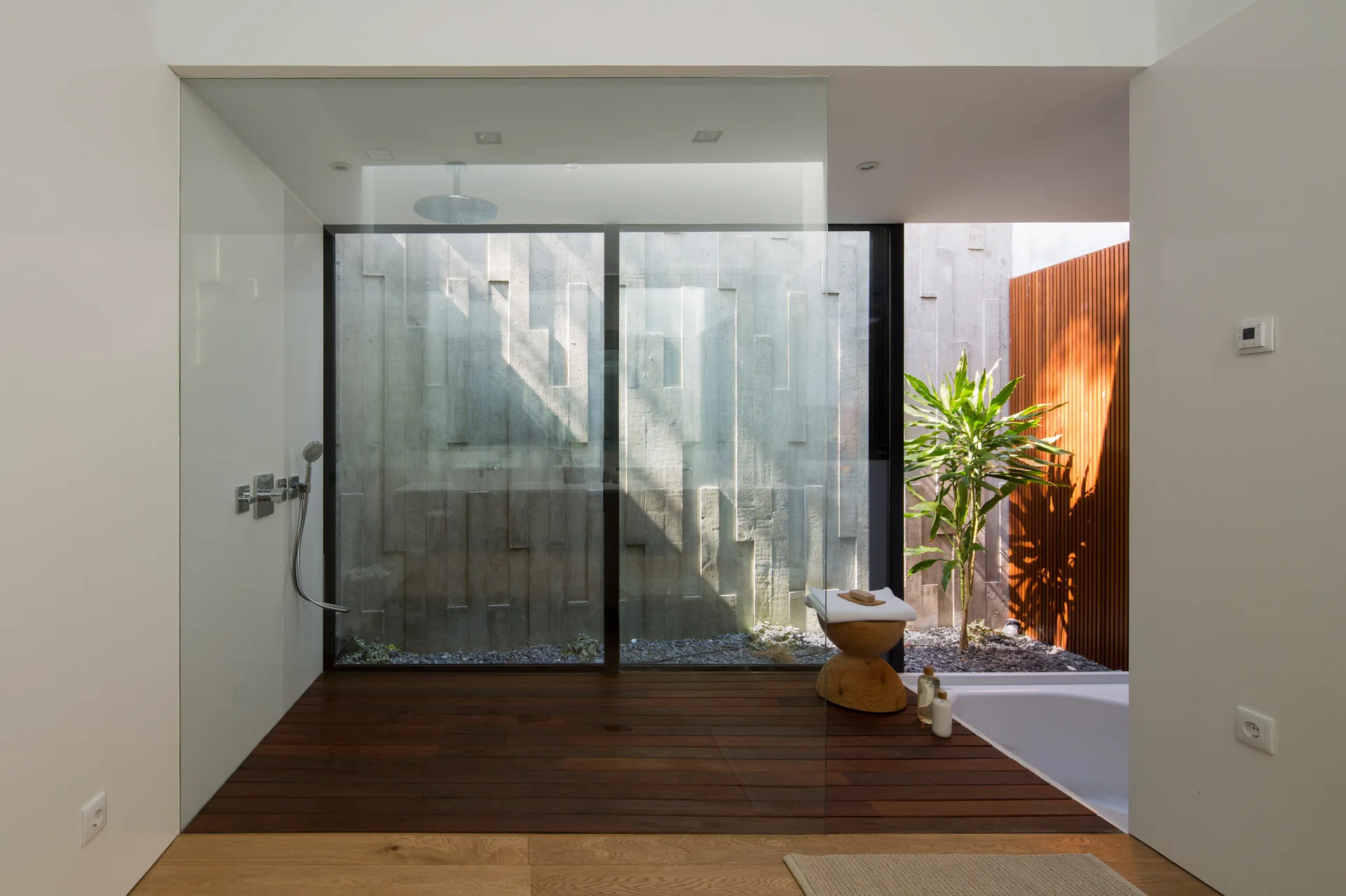 A minimalist bathroom with dark wood plank flooring, a frameless glass partition, a white bathtub, and a view into a concrete lightwell.