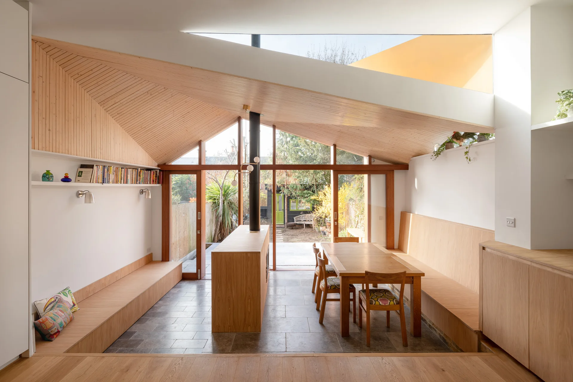 A sunlit dining room features a wood-slatted vaulted ceiling, a long wooden dining table, and built-in benches next to glass garden doors.