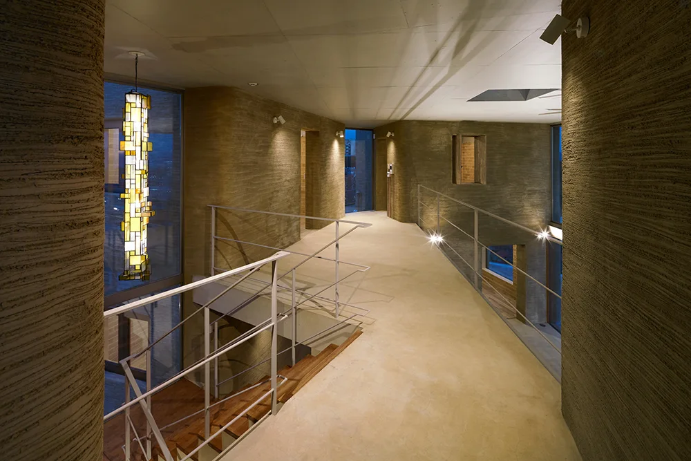An interior elevated walkway features heavily textured earth walls, slim white metal railings, and a glowing yellow pendant light.