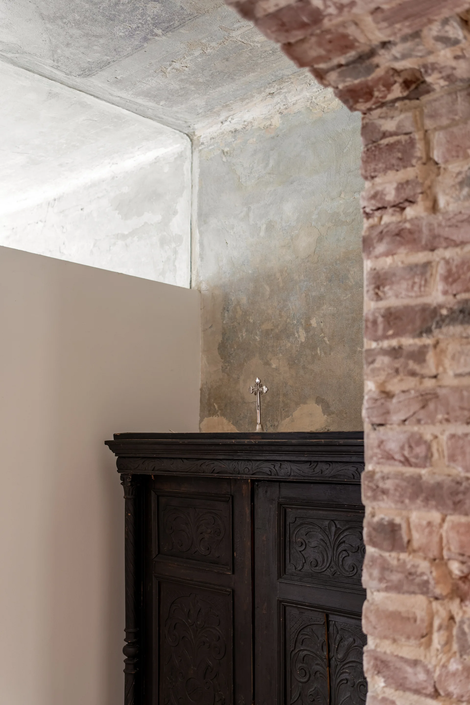 An ornate dark carved wood cabinet stands against a highly distressed grey plaster wall beside an exposed red brick corner.