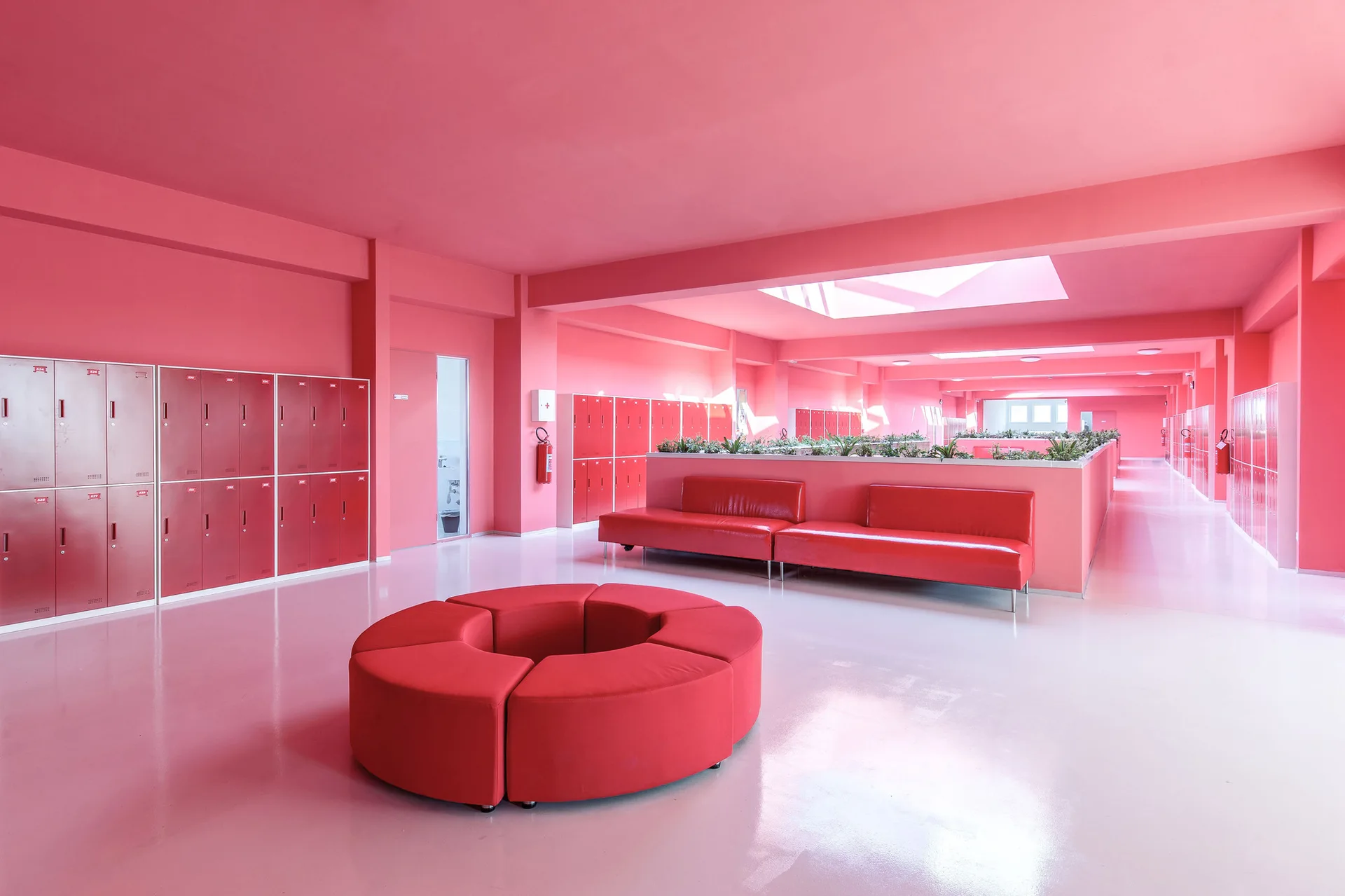 A large monochromatic pink institutional lobby features a circular red modular ottoman, red sofas, and matching red metal lockers.