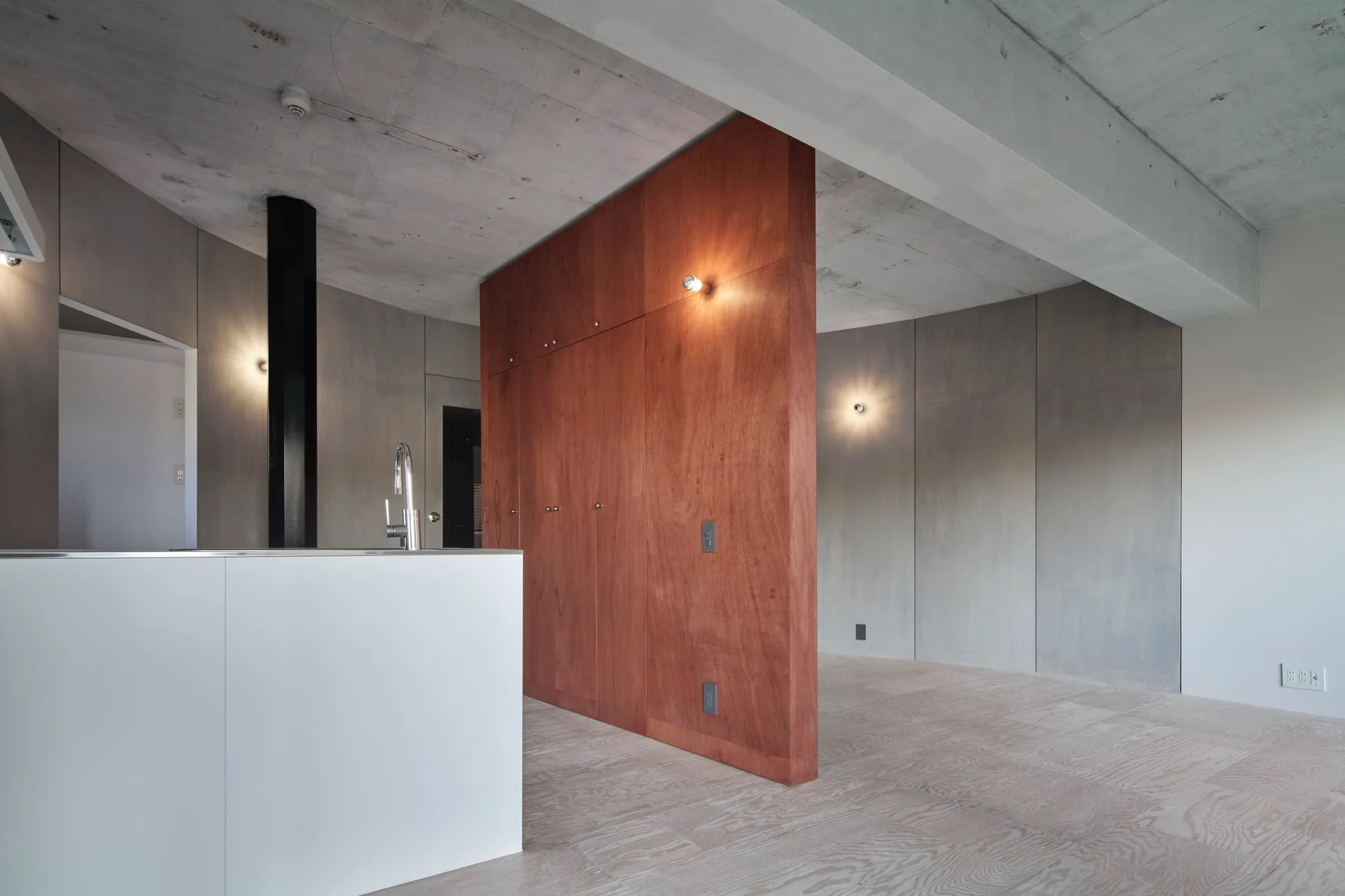 A minimalist kitchen featuring a white island and a prominent reddish-brown wood partition wall set against an exposed concrete ceiling.