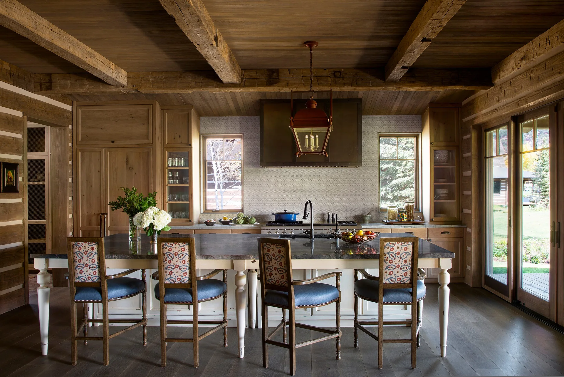 A rustic kitchen features a large wood island with a stone countertop, surrounded by four patterned bar stools beneath an exposed beam ceiling.