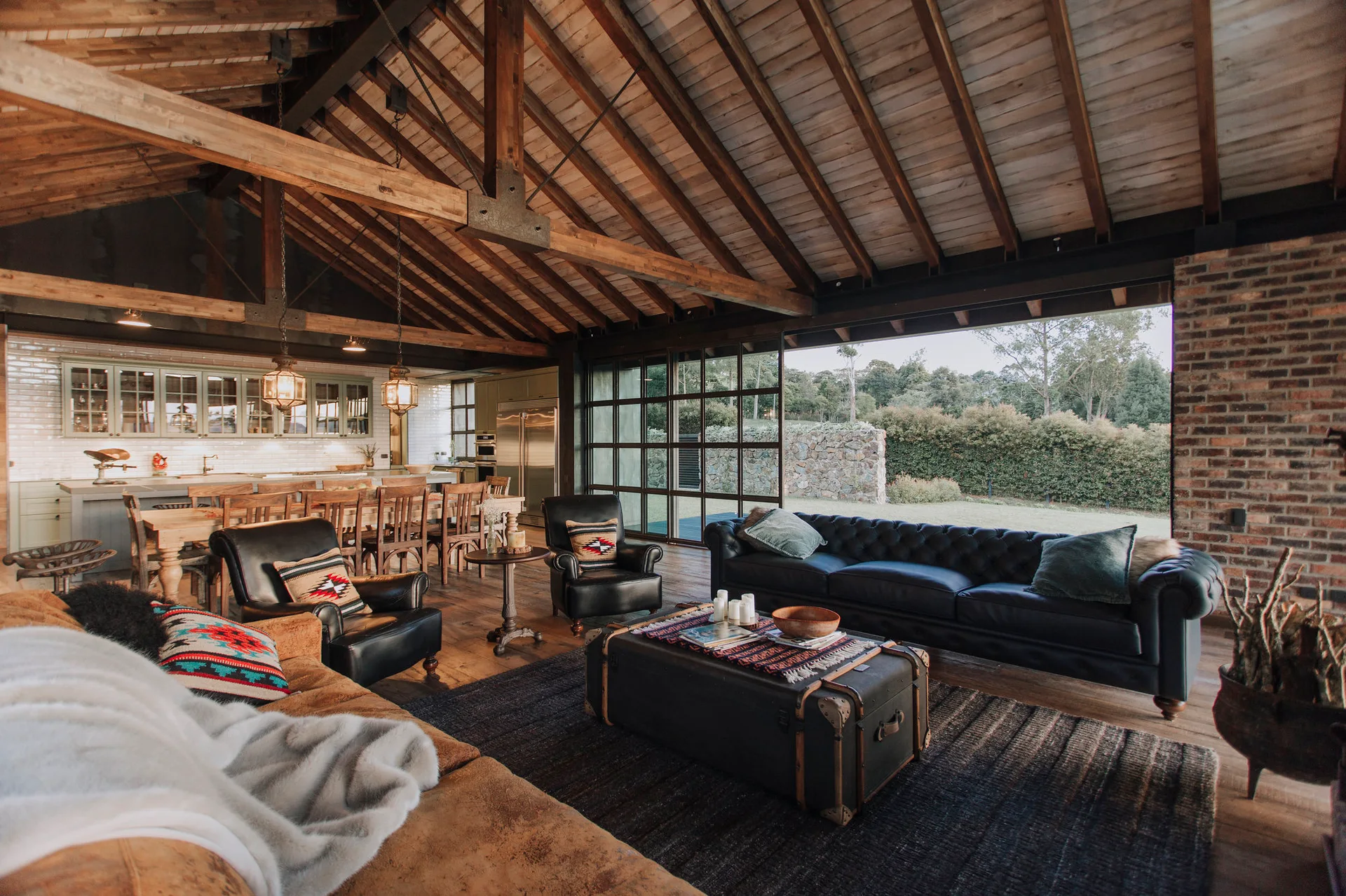 A rustic living room features a vaulted wood ceiling, black leather sofas, and a large black-framed glass wall opening to the outdoors.
