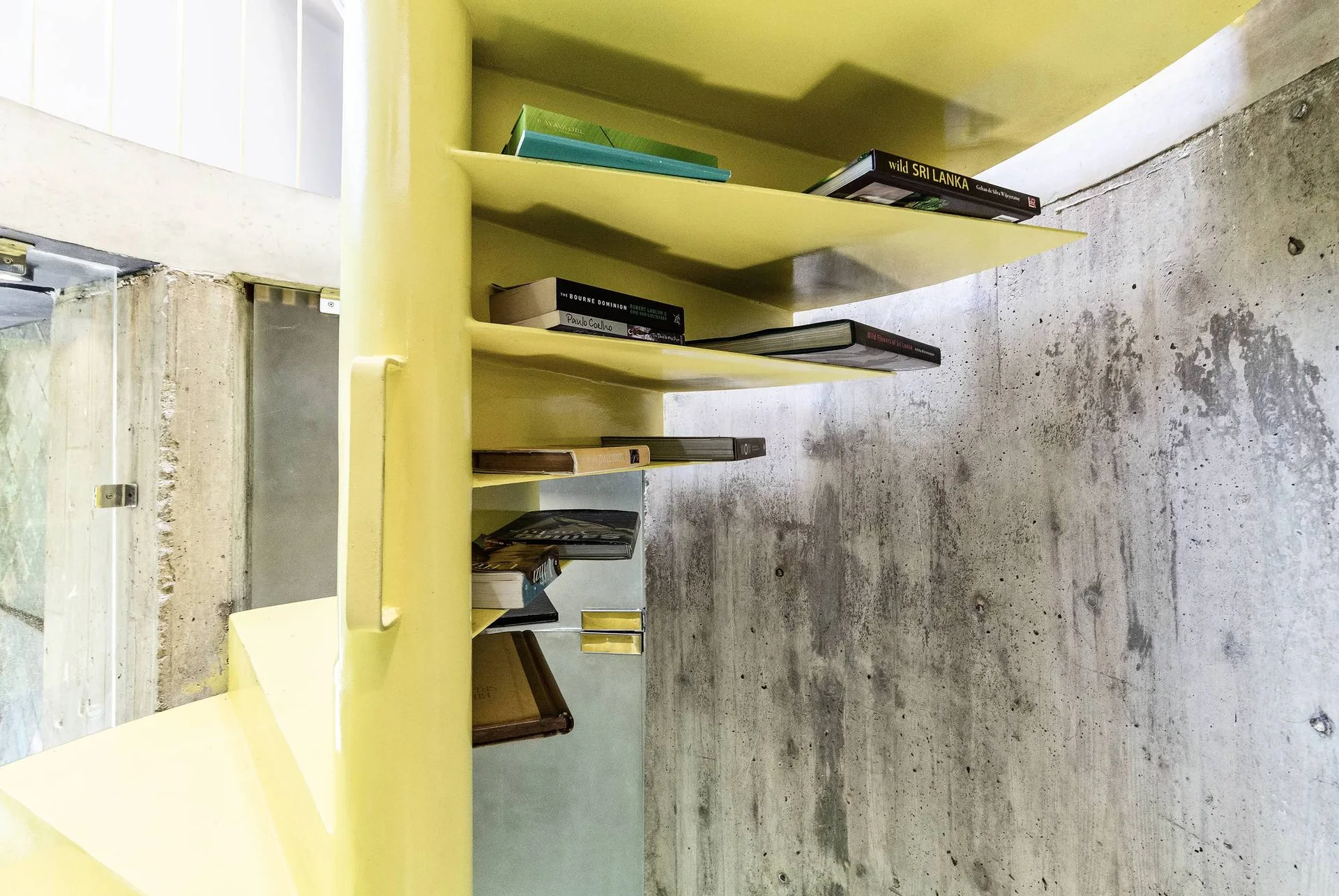 A yellow metal spiral staircase with integrated book shelves is positioned against a raw textured gray concrete wall.