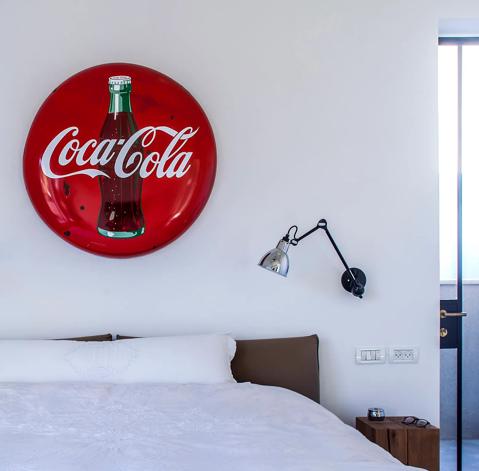 A minimalist bedroom featuring a large red Coca-Cola sign mounted above a bed with white bedding, paired with a black adjustable swing-arm wall sconce.