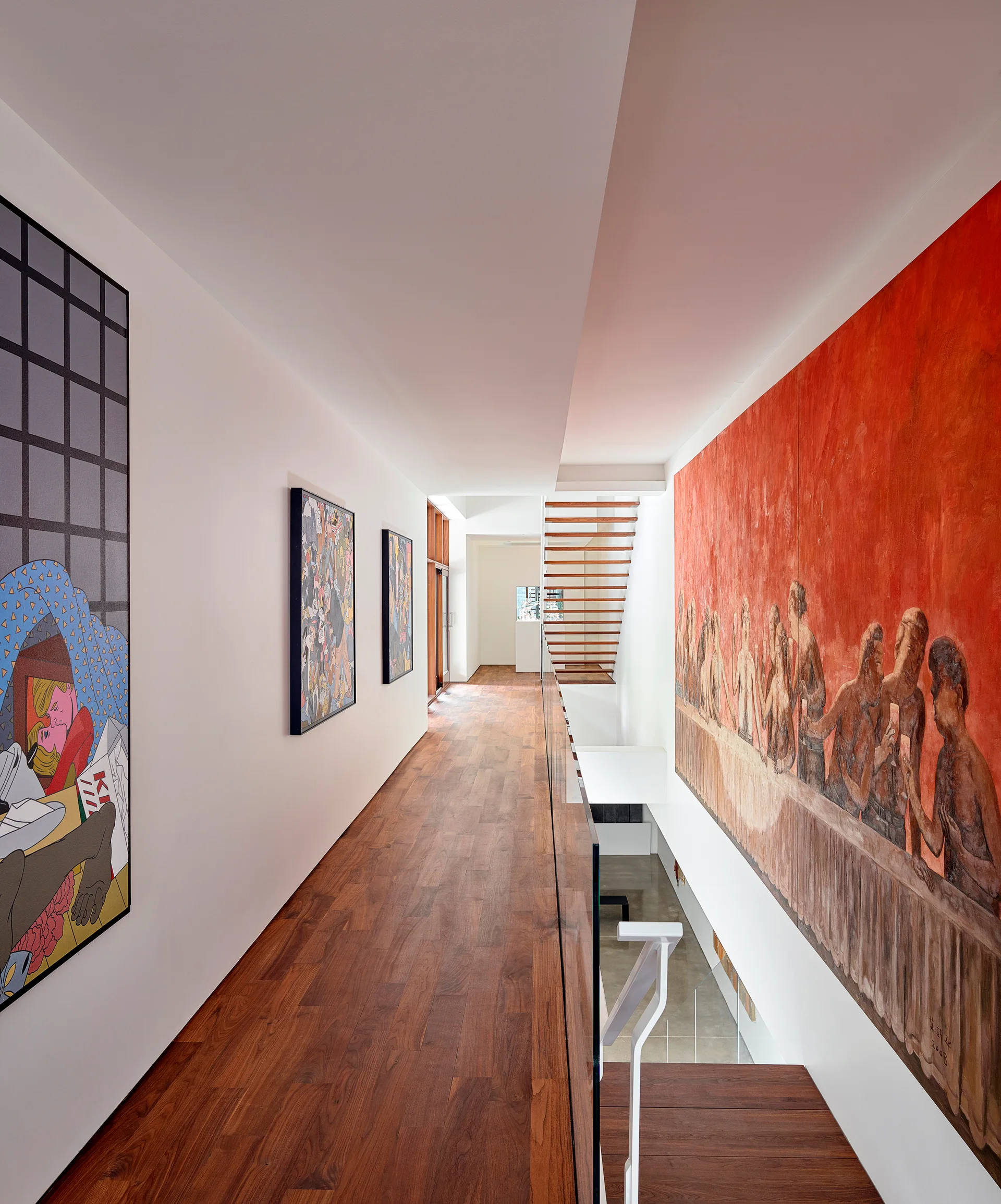 A long contemporary hallway with dark wood flooring features white walls hung with colorful art and a glass railing overlooking an open staircase.