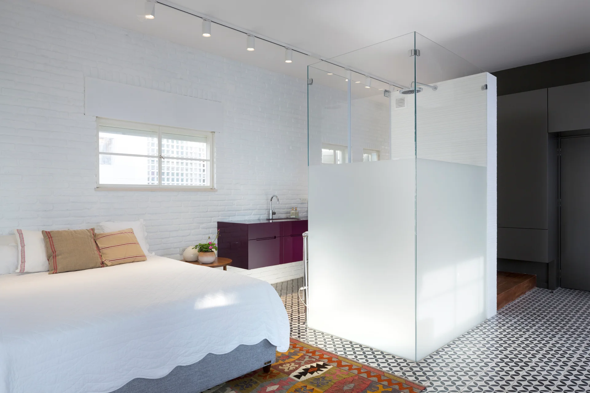 A modern open-concept bedroom featuring a glass-enclosed shower, a white painted brick wall, a floating purple vanity, and geometric floor tile.