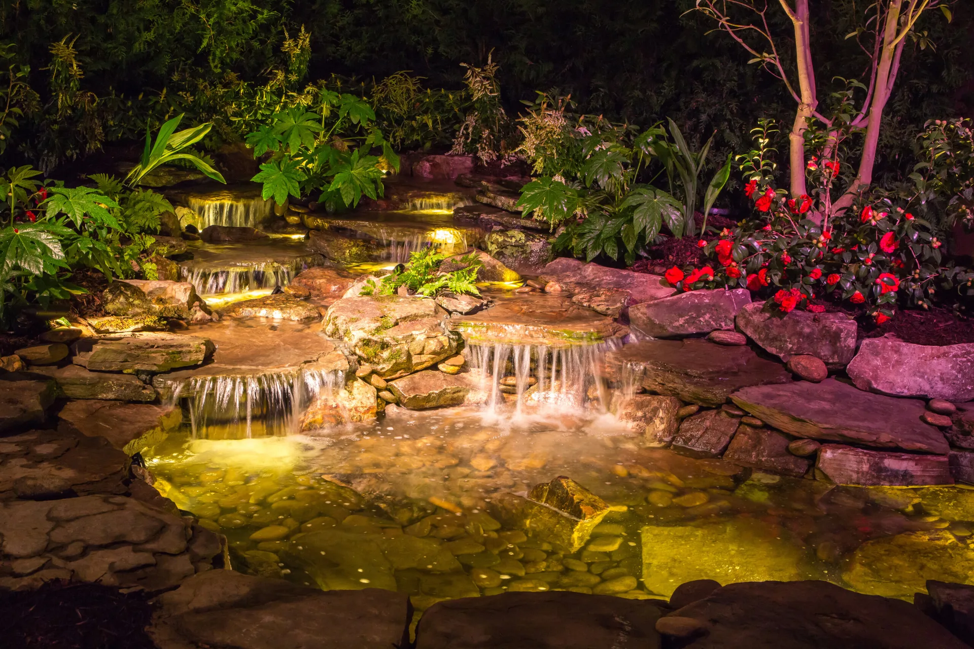 A tiered stone waterfall flows into an illuminated pond at night, surrounded by lush green foliage and red flowering shrubs.