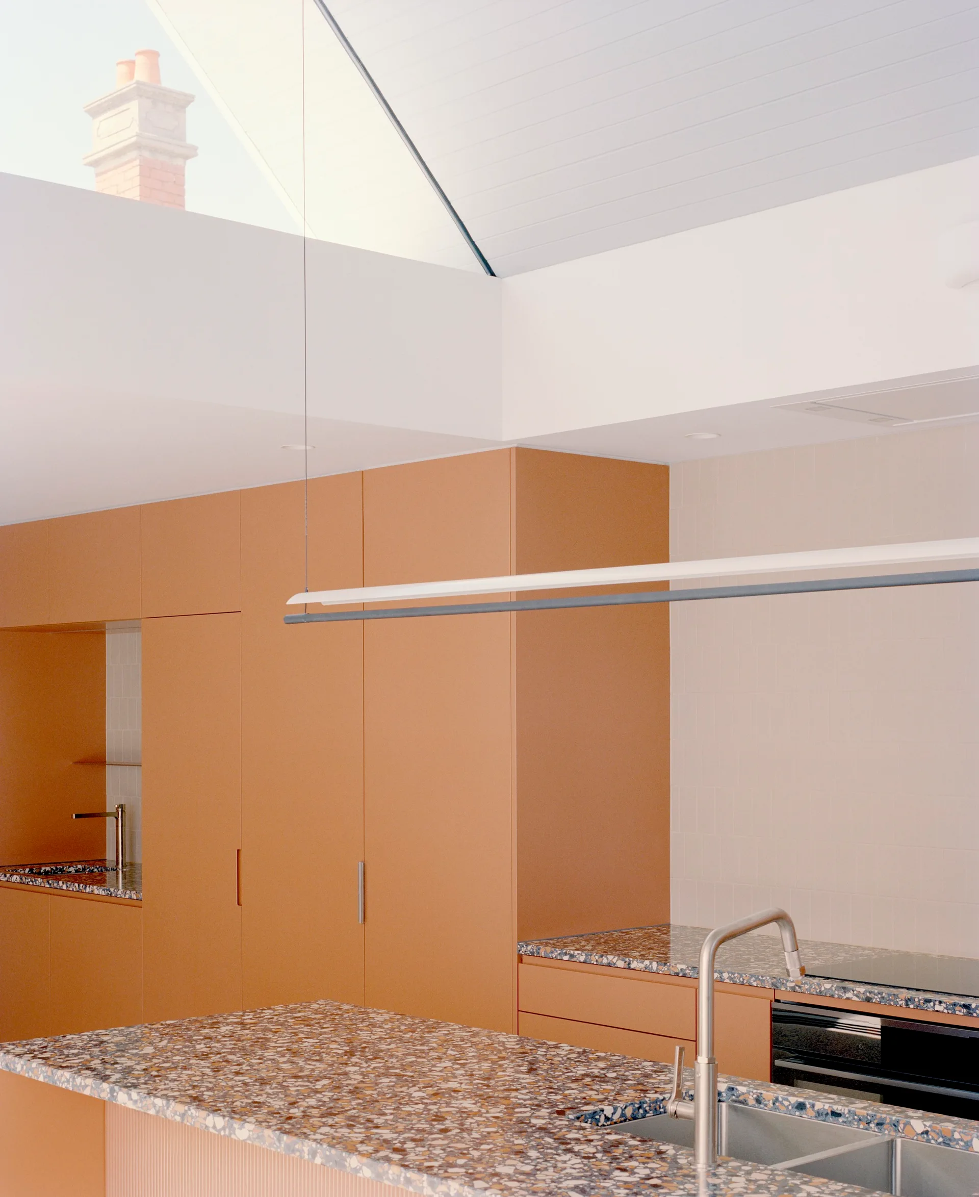 A modern kitchen featuring matte terracotta-orange cabinets, a terrazzo-topped island with a stainless steel sink, and light streaming from a high skylight.