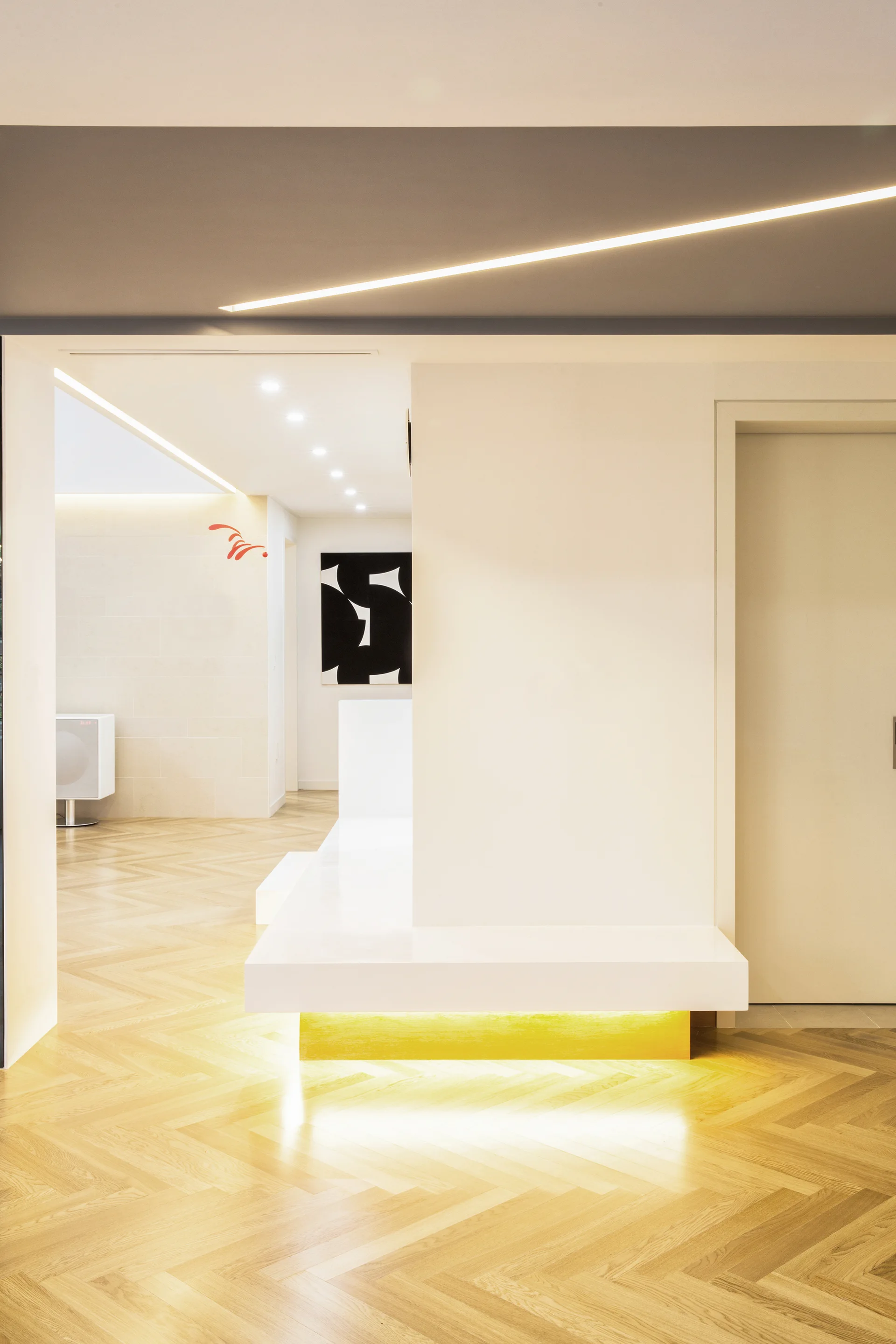 A modern hallway features light herringbone wood flooring, a built-in white bench with warm under-lighting, and recessed linear ceiling lights.