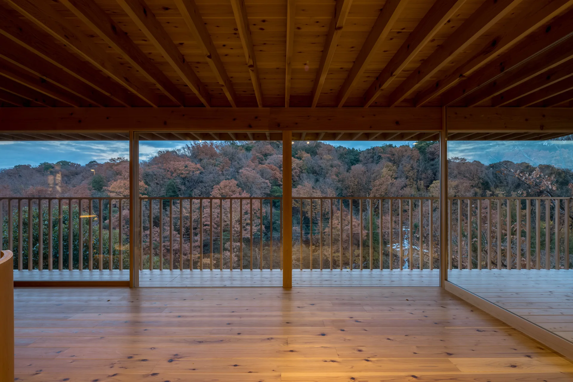 A minimalist room features a warm wood floor and exposed ceiling beams with large glass windows showing a balcony and autumn forest.