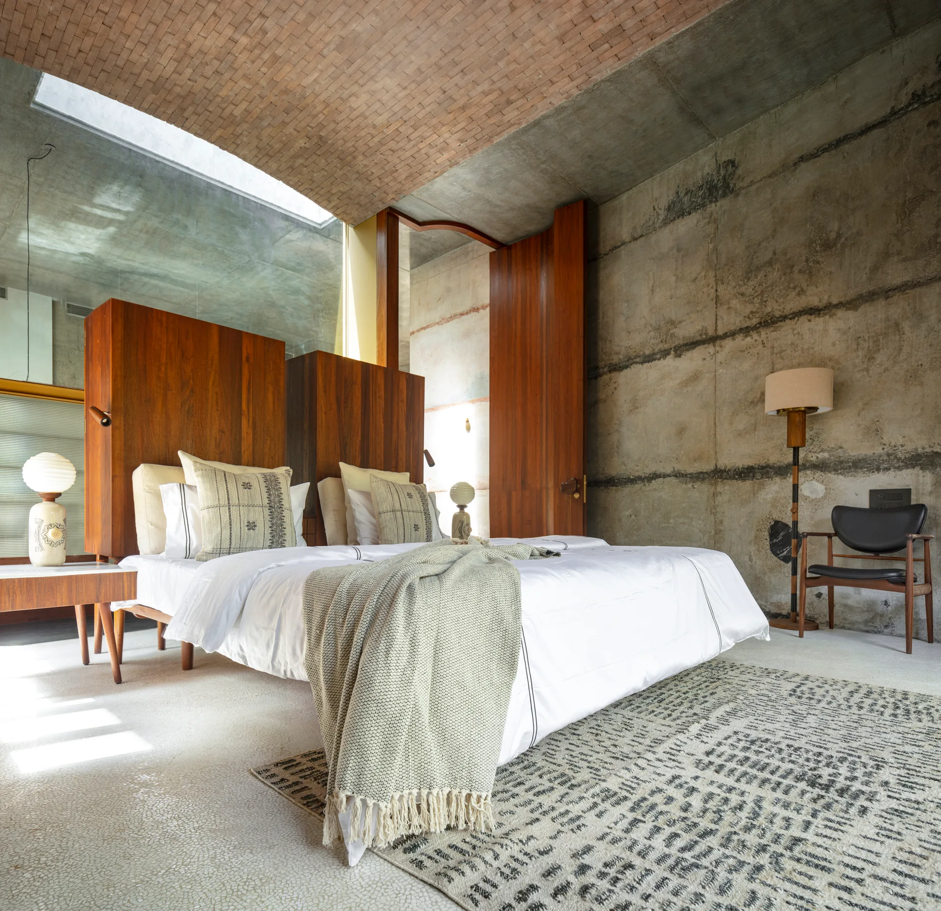 A bedroom features raw concrete walls, a vaulted brick ceiling with a skylight, a wood panel headboard, and a white bed on a patterned rug.