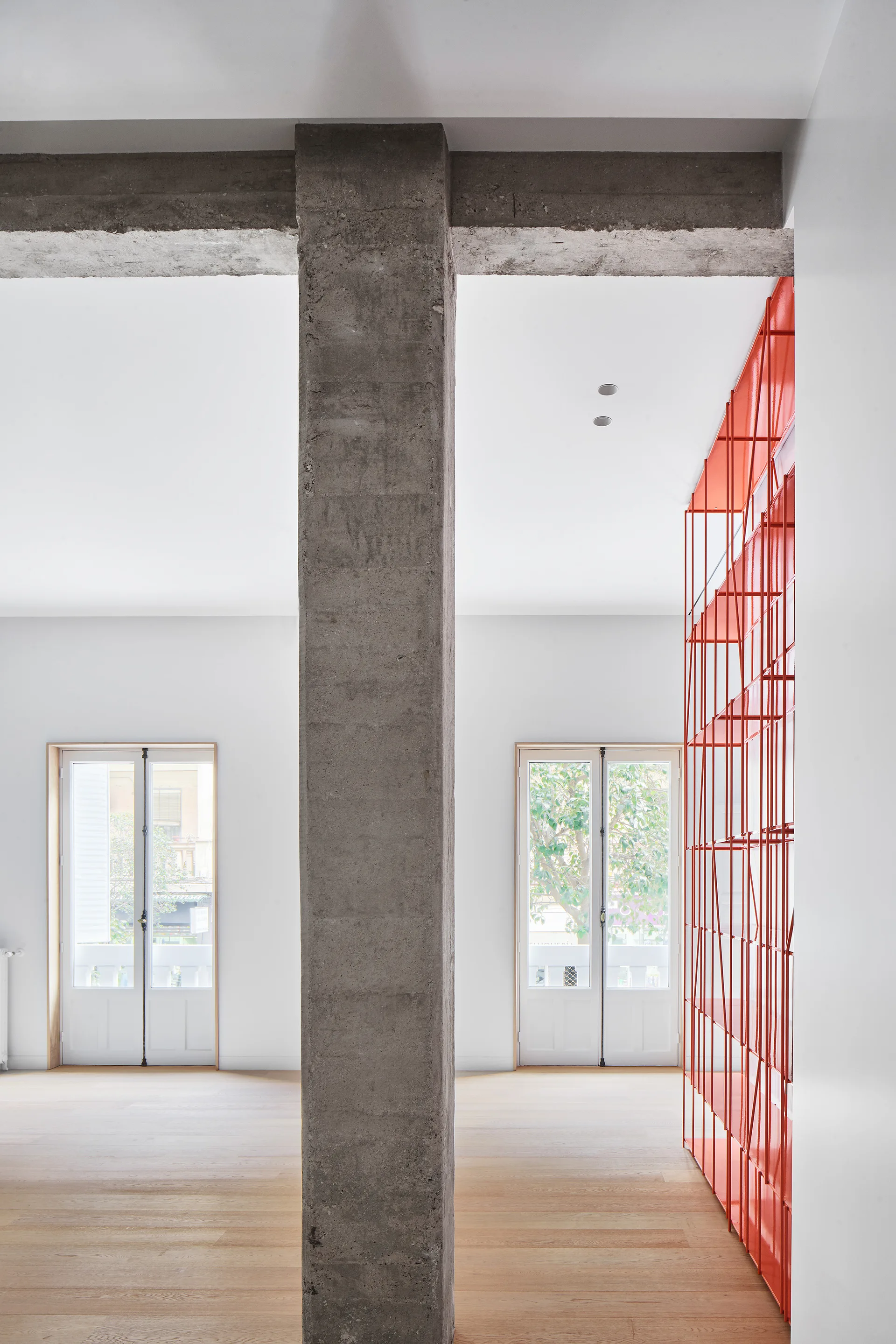 An airy minimalist interior features a central concrete column, light wood flooring, white walls, and a tall red metal shelving unit.