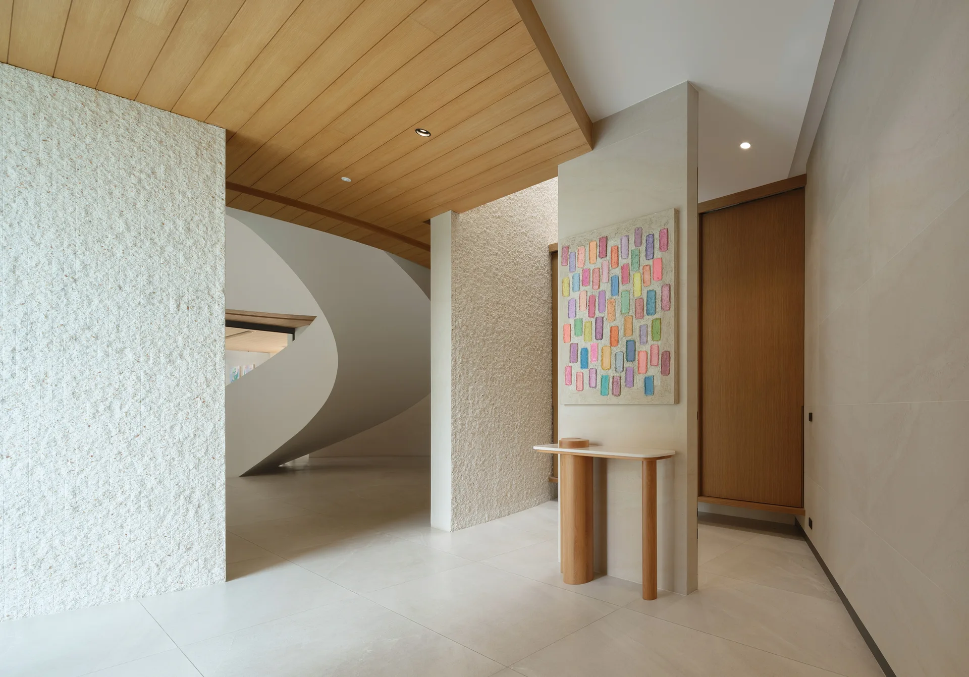 An organic modern foyer features textured stone walls, a slim wooden console table with colorful art, and a curved sculptural staircase.