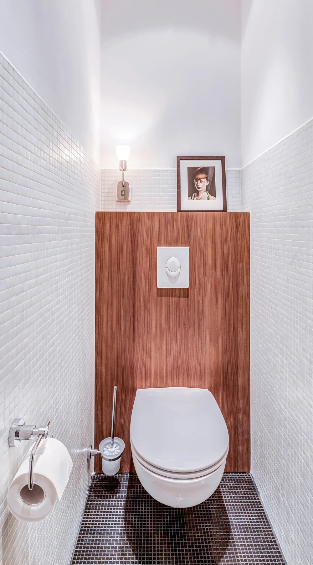 A small powder room with a white wall-mounted toilet against a vertical wood panel, white mosaic wall tiles, and black tiled flooring.