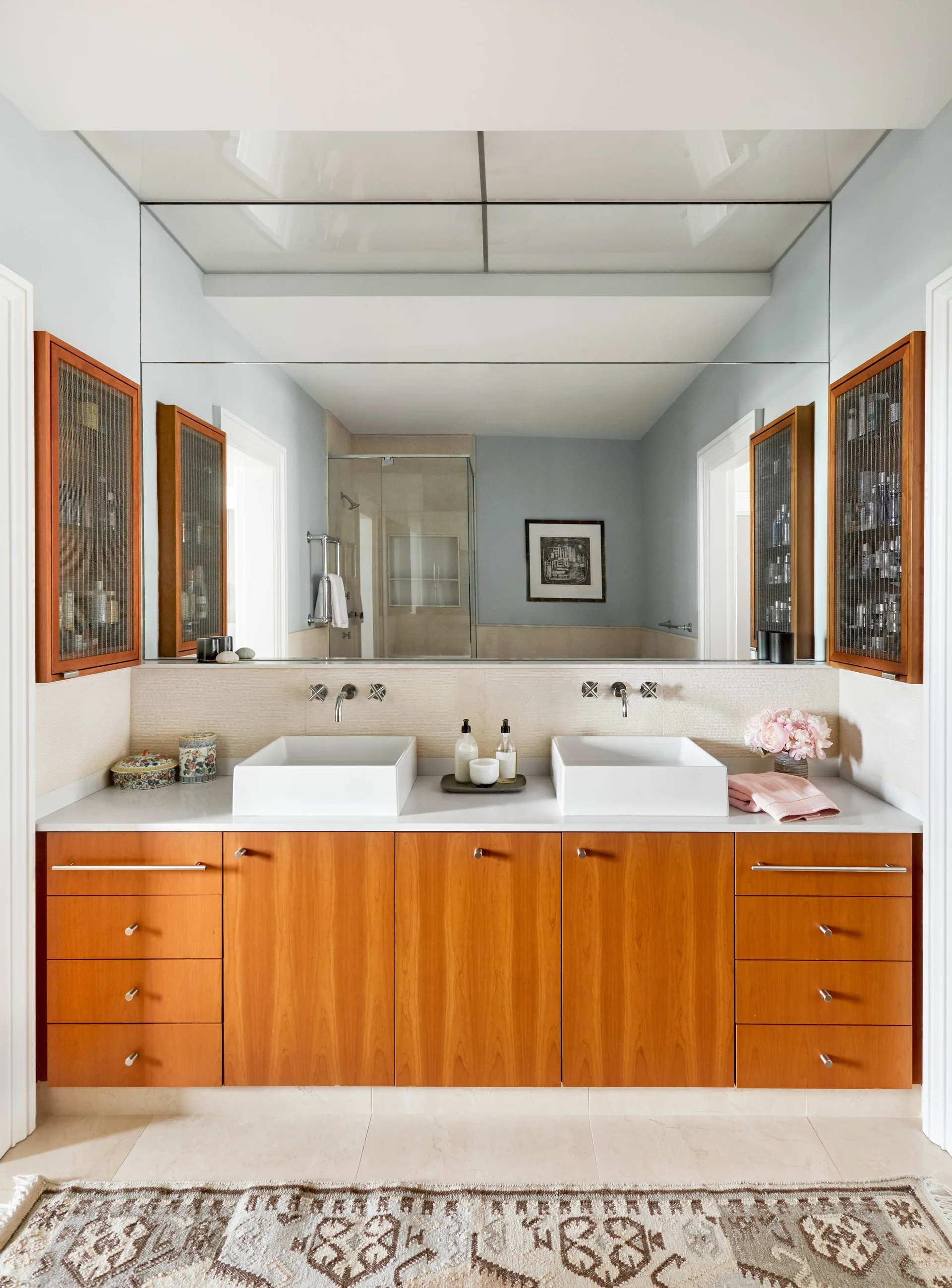 A contemporary bathroom vanity with warm wood cabinets, two white vessel sinks with wall-mounted faucets, and side cabinets with wire mesh doors.