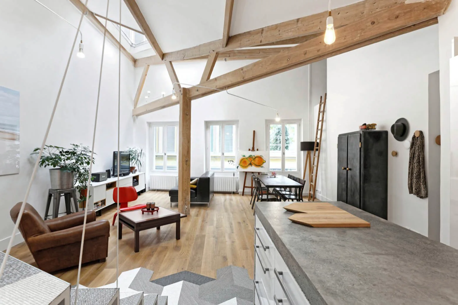 An open-plan loft living space featuring exposed wooden ceiling beams, light hardwood floors, a brown leather armchair, and a concrete-top kitchen island.