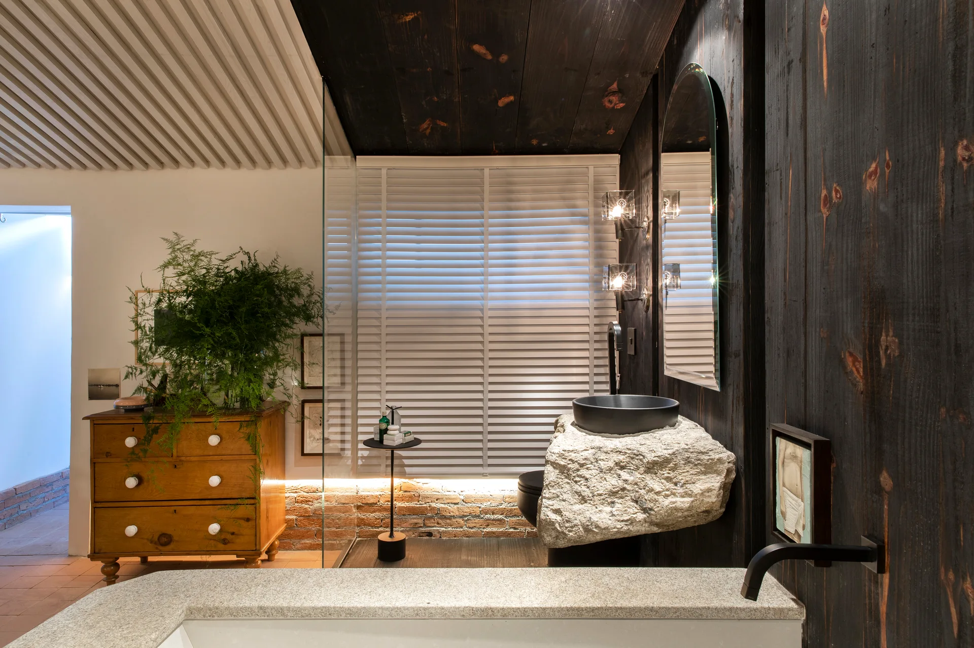 A modern bathroom features a sleek black vessel sink mounted on a rough natural stone block against charred wood wall panels.