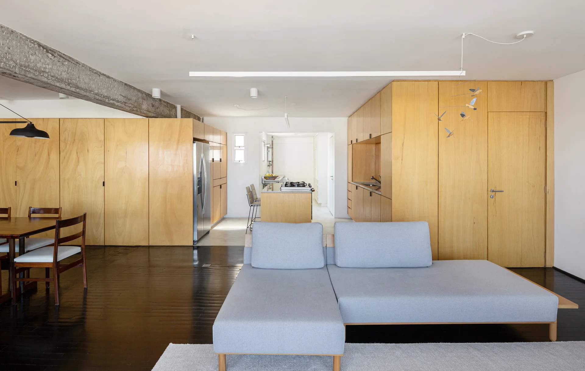 A light grey sectional sofa on dark wood floors in an open-plan living room featuring extensive light plywood cabinetry and an exposed concrete ceiling beam.
