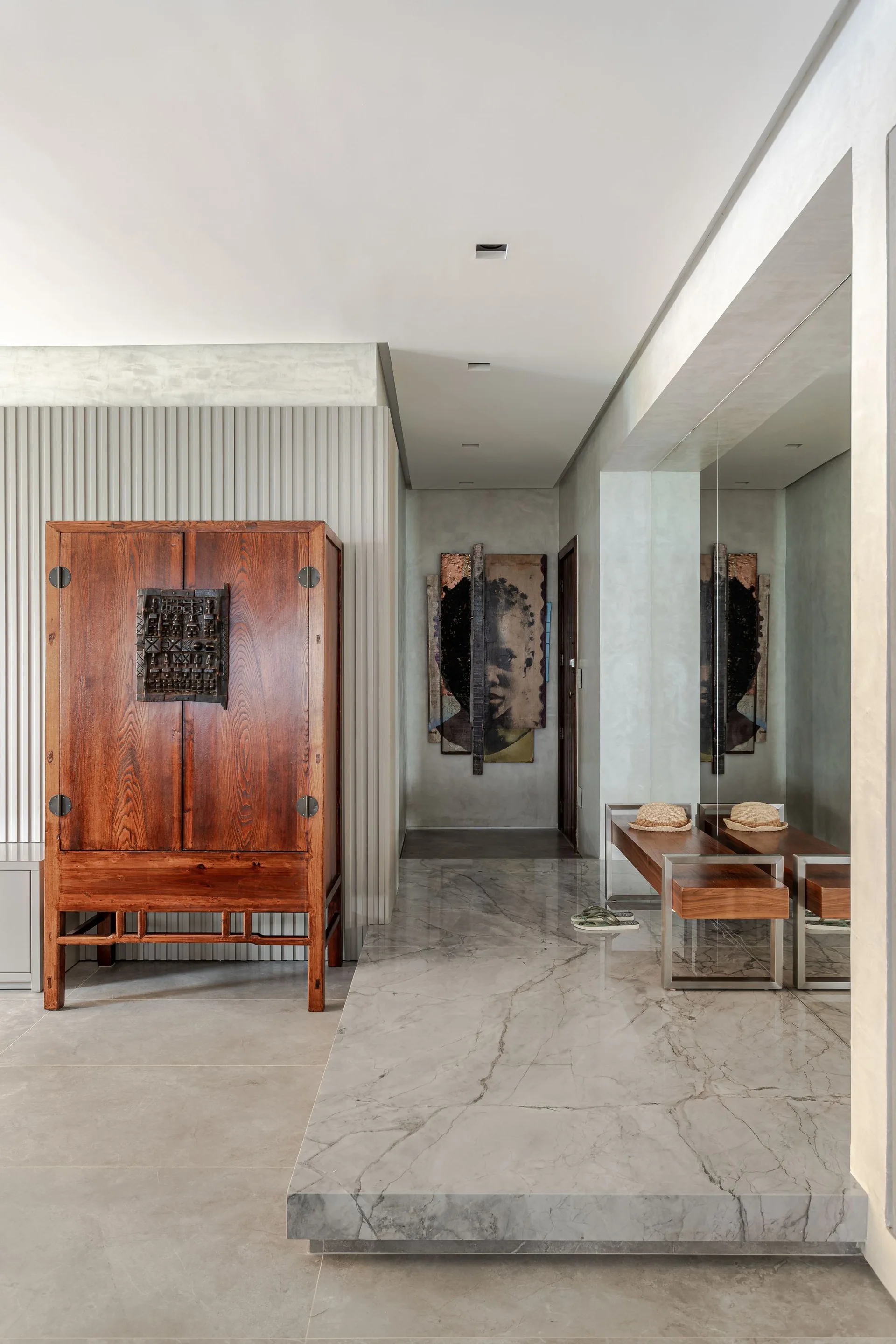 A modern entryway featuring a tall wooden cabinet against a ribbed wall, a marble platform floor, and a wooden bench in front of a mirrored wall.